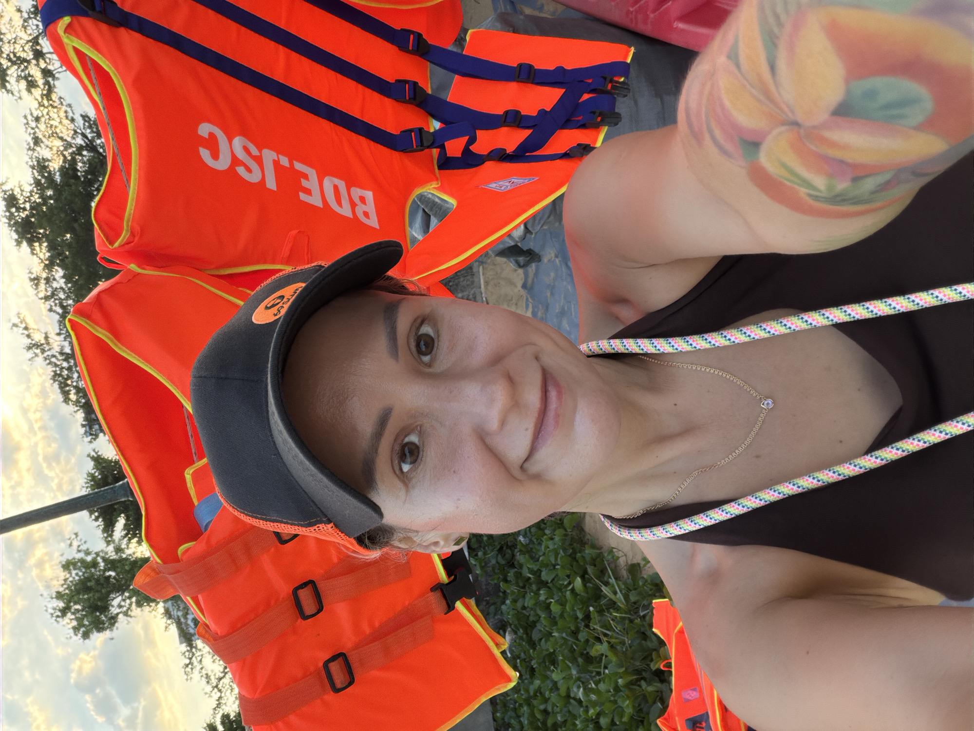 A woman with a tattoo on her right arm and wearing a black baseball cap and a dark brown halter top takes a selfie outdoors. Behind her, several bright orange life vests are stacked. The sky, with scattered clouds, is visible in the background. The scene appears to be in the city of South Quy Nhon Ward, Vietnam, near a grassy area.