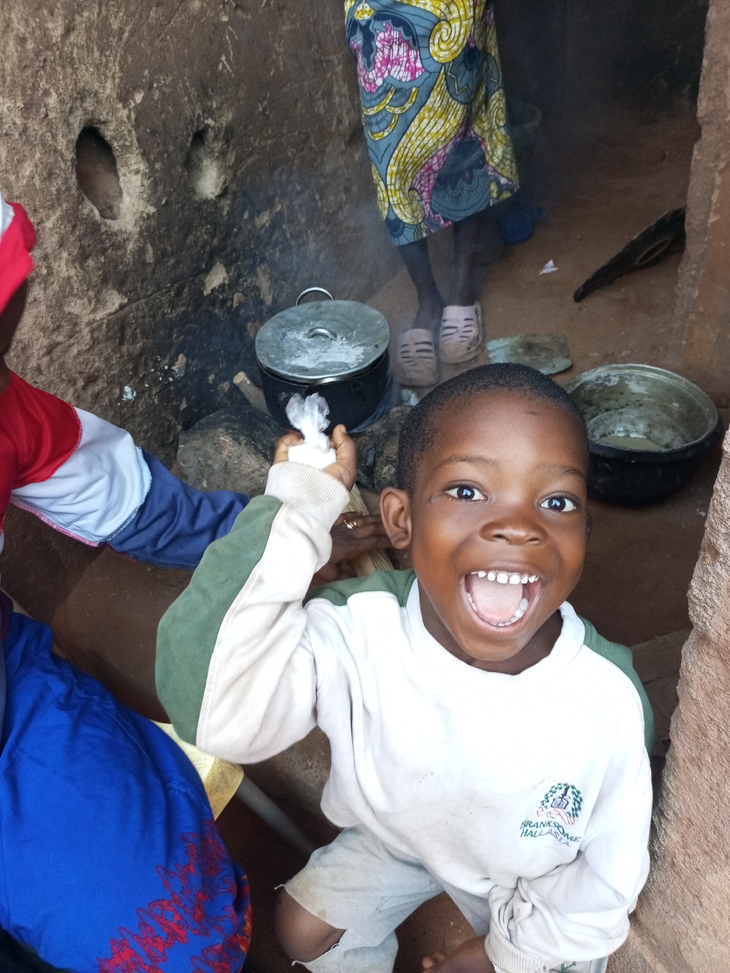 A boy grins broadly with his mouth open while holding up a small white object in his left hand. He wears a white long-sleeved sweatshirt with green accents and light-colored shorts. Behind him, to his left, an adult wearing a colorful patterned skirt stands near a cooking pot on a fire. To the boy's right, another pot sits on the ground. The scene appears to be outdoors, possibly in a rural area of Tempure, Nigeria, with rough, textured walls forming the background and an earthen floor.