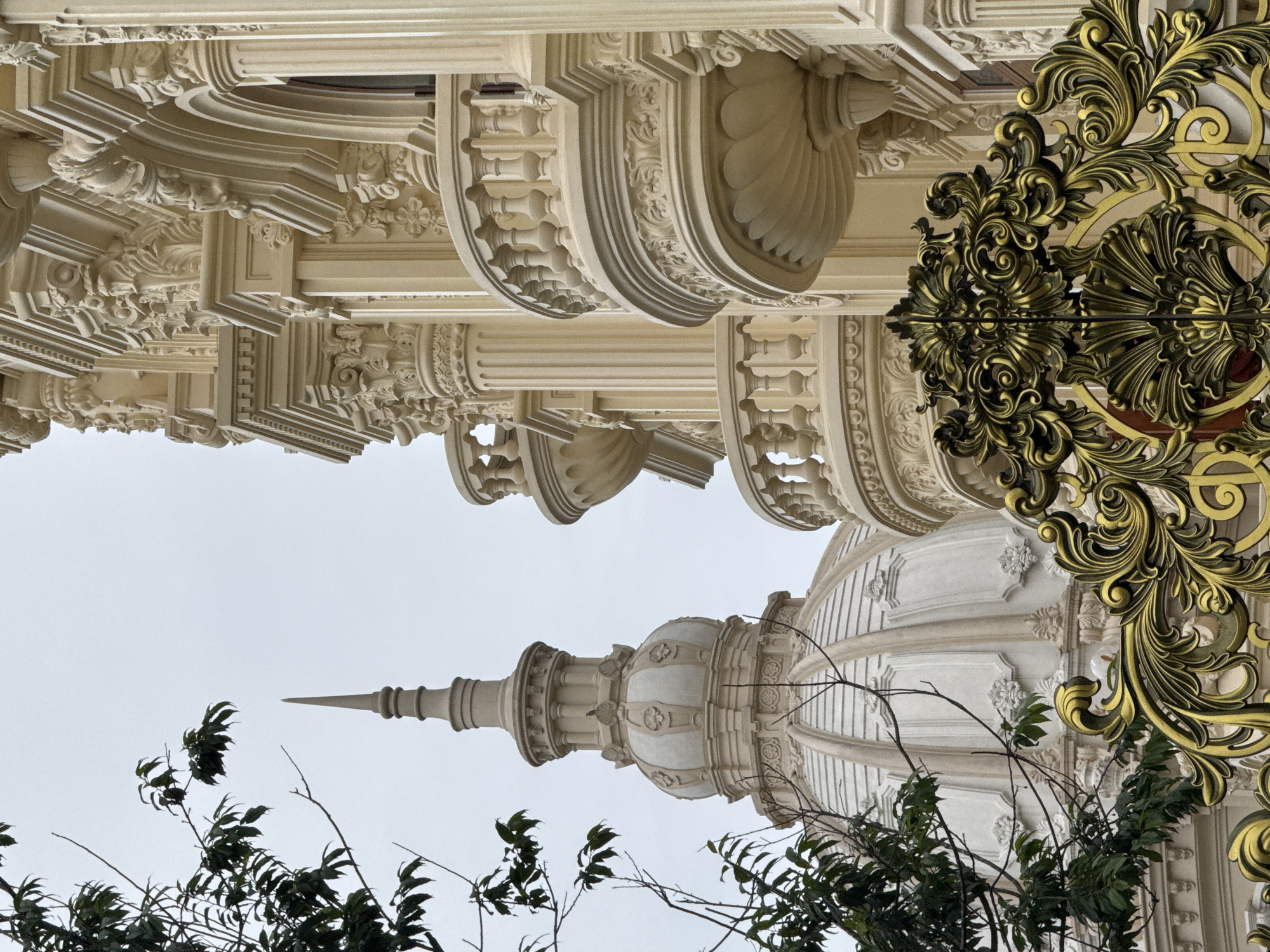 The image captures a close-up view of an ornate building with neoclassical architectural elements, likely located in Da Lat, Vietnam. In the foreground, a portion of a decorative golden gate with intricate scrollwork is visible. The building itself features a dome structure with a spire, partially obscured by dark green foliage from a tree on the left. The main facade of the building, to the right, is adorned with detailed carvings, columns, and balconies with balustrades, all rendered in a light cream color. The sky is overcast and light grey. The scene is static, with no people or discernible activity.