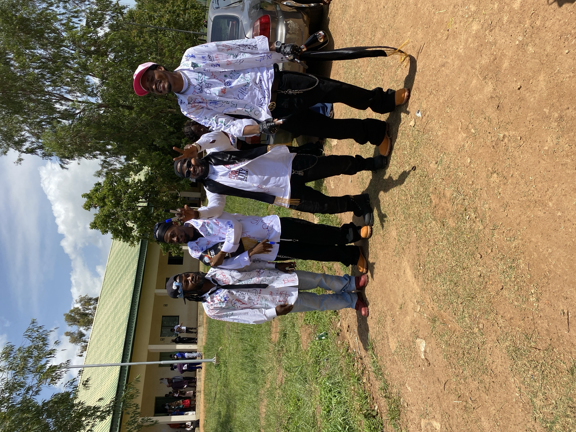 Four young men are posing for a photograph outdoors, in front of a light-colored building with a green roof. The building appears to be an educational institution, with a row of windows and people gathered in the background. The ground in the foreground is a dirt and grass mixture. The men are all wearing white shirts covered in colorful inscriptions and drawings, likely signatures or messages from a celebration or graduation event. They are dressed in casual streetwear. One man on the far left wears a black bandana and jeans. The next man to his right has on a black vest and sunglasses. The third man, on the right side of the frame, is wearing a pink baseball cap. The fourth man, on the far right, is also wearing a white shirt and black pants. A car is partially visible behind them to the right. The sky is blue with scattered white clouds. The location is identified as Wumat, Nigeria.