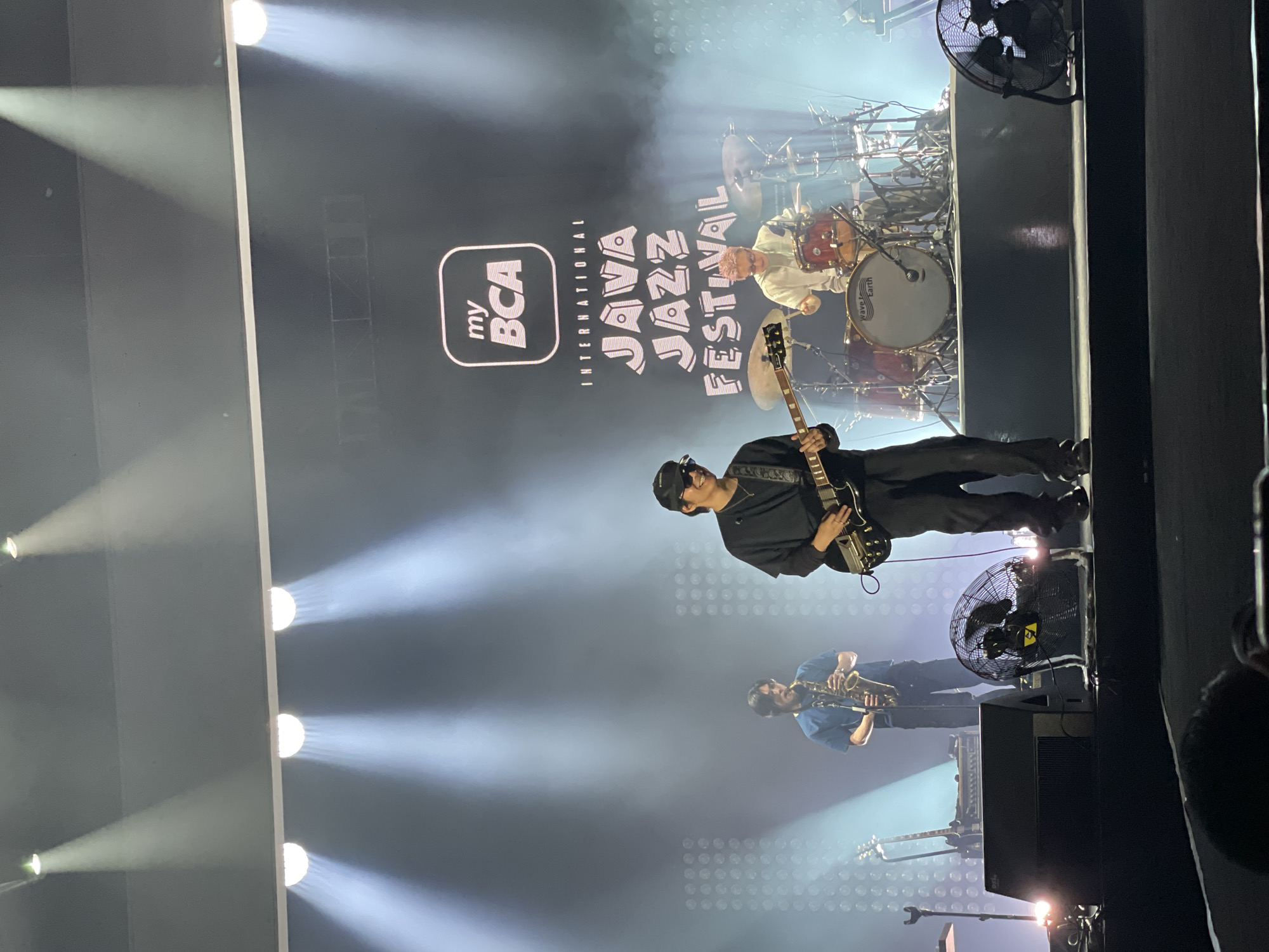 The image captures a live performance at the my BCA International Java Jazz Festival in Salembaran, Indonesia. The stage is lit with spotlights, creating a bright atmosphere. A guitarist, dressed in all black, stands center stage. Below him, a saxophone player in a blue shirt is seen. Behind the guitarist, a drummer is visible behind his drum kit. There is a logo for the festival, "my BCA International Java Jazz Festival," on a screen behind the musicians. A large electric fan sits on the stage as well. The scene conveys the ambiance of a live music event.