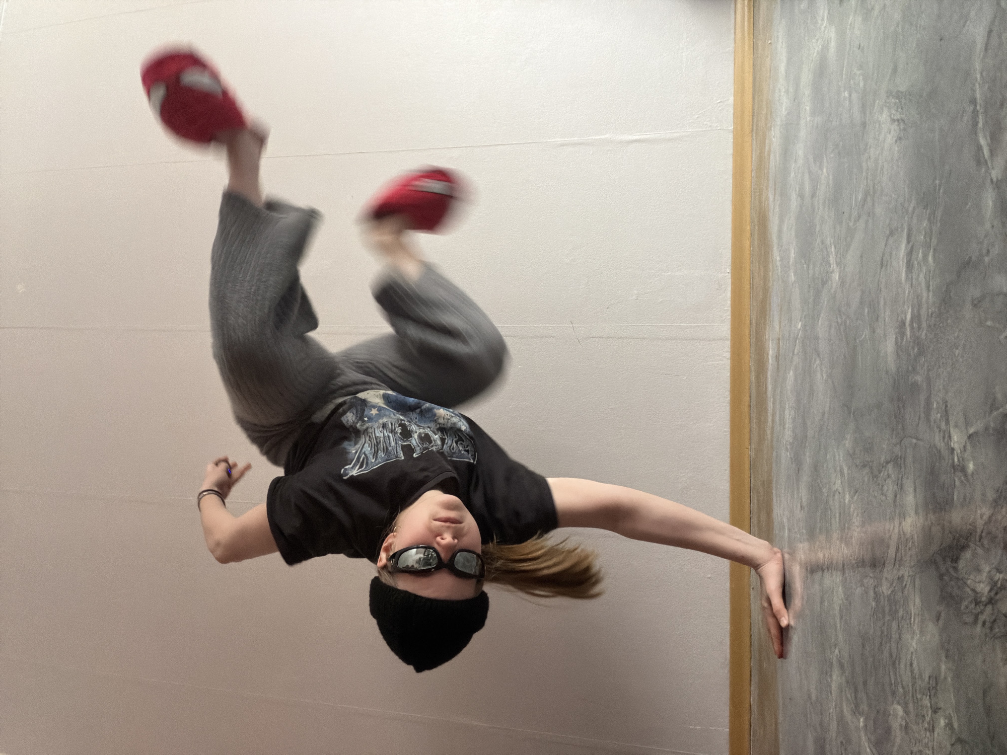 A person is performing an acrobatic handstand against a white wall. The person is wearing a black beanie, sunglasses, a black t-shirt, gray sweatpants, and red slippers. The person is leaning against a gray textured wall with one hand. The photo was taken in Perm, Russia.