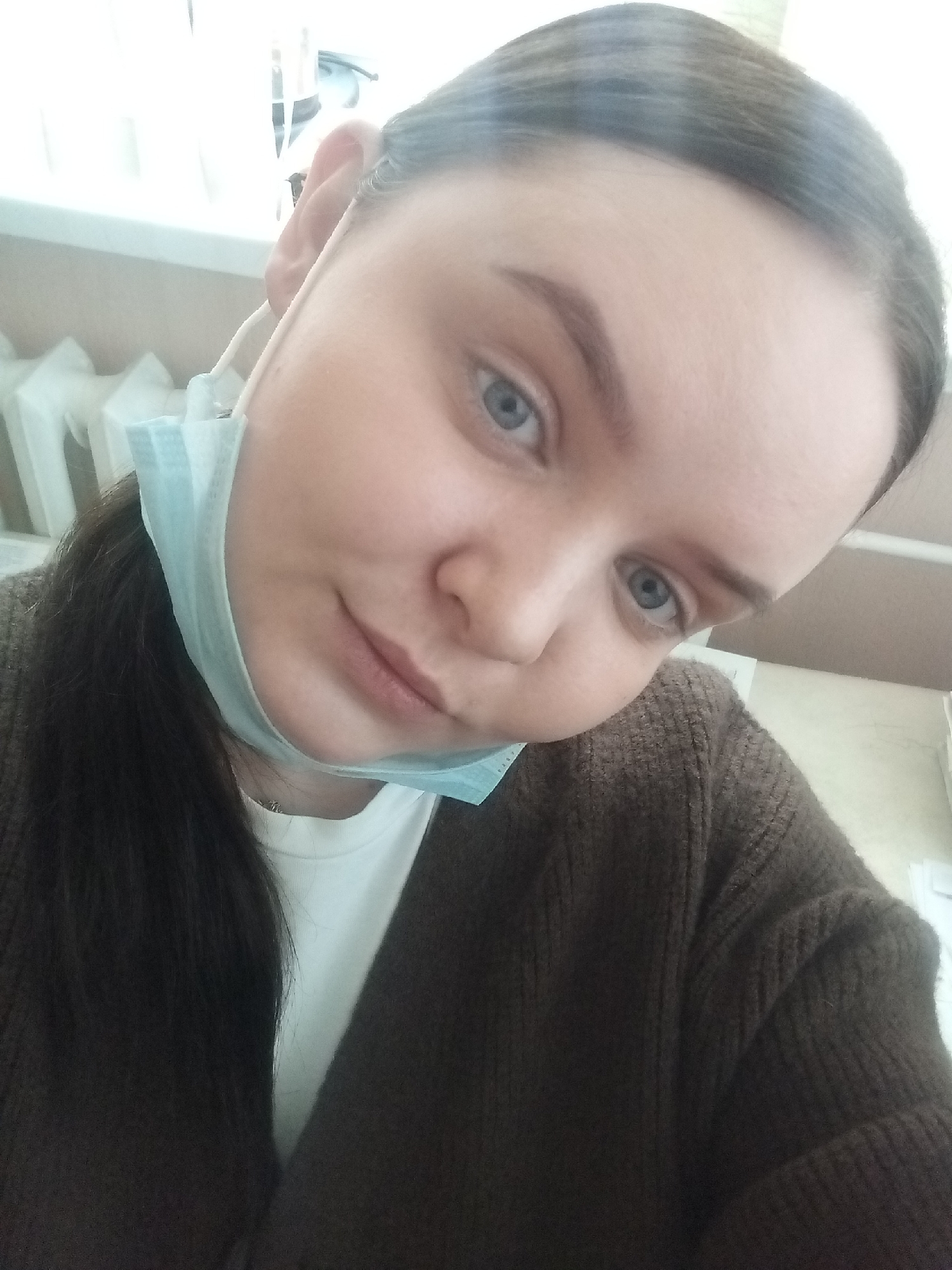 A close-up, slightly upward-angled portrait shot in an indoor setting in Chaikovsky, Russia, depicts a young adult female. She has fair skin, blue eyes, and dark eyebrows. Her dark hair is pulled back into a ponytail, visible over her left shoulder. She wears a brown, ribbed knit cardigan over a white top. A light blue surgical-style face mask is present, pulled down to rest beneath her chin, exposing her mouth and nose. The mask's ear loop is visible around her left ear. Her gaze is directed towards the camera, and her mouth forms a subtle, closed-lip smile. The background on the left features a brightly lit window with a white frame and horizontal blinds, and a white radiator visible below. The background on the right shows light-colored walls and some indistinct white objects.