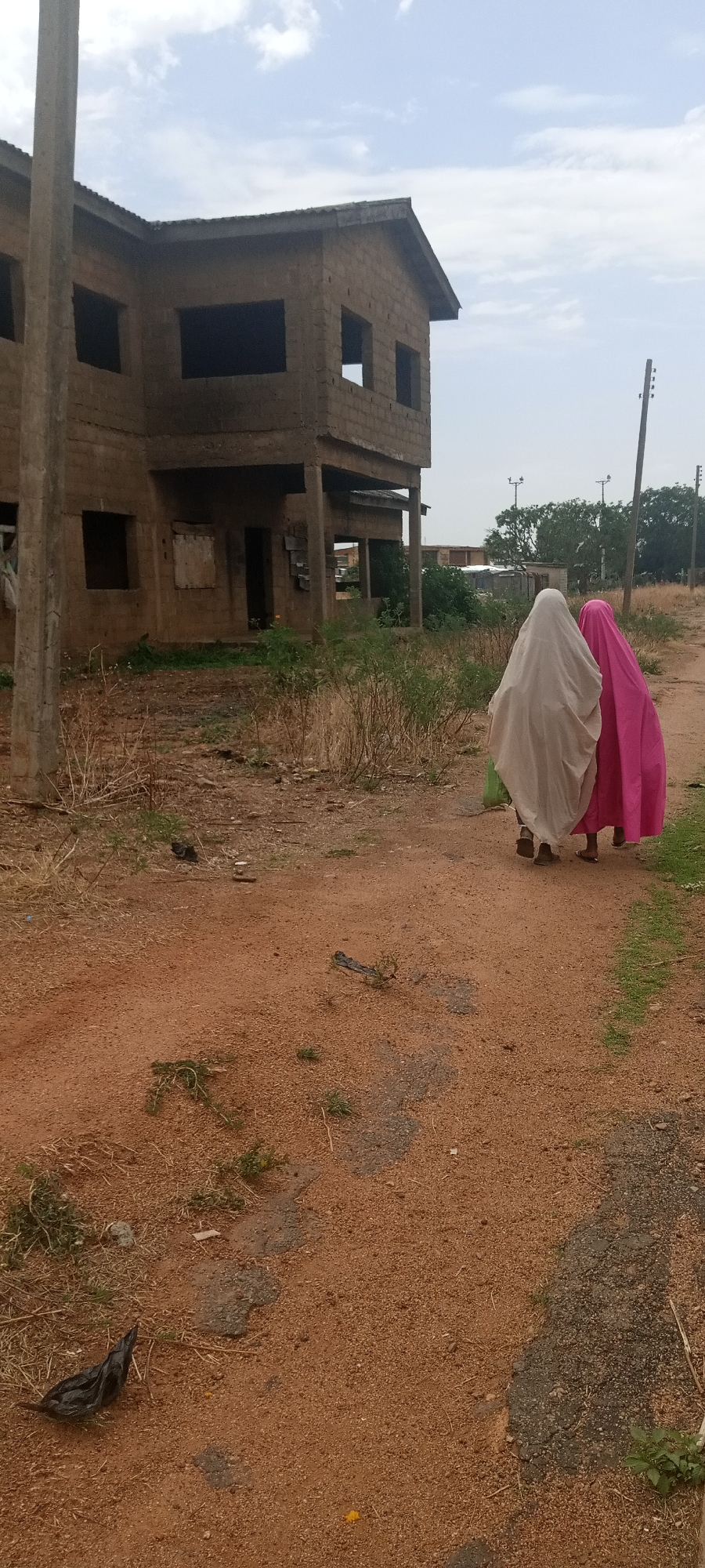 Two women are walking along a reddish dirt path, dressed in full-body coverings, one light grey and the other pink. To their left is a partially constructed, two-story brick building with open window frames. The path is littered with sparse vegetation and a black plastic bag in the foreground. In the background, utility poles and trees are visible. The location is in the city of Unknown, Unknown.
