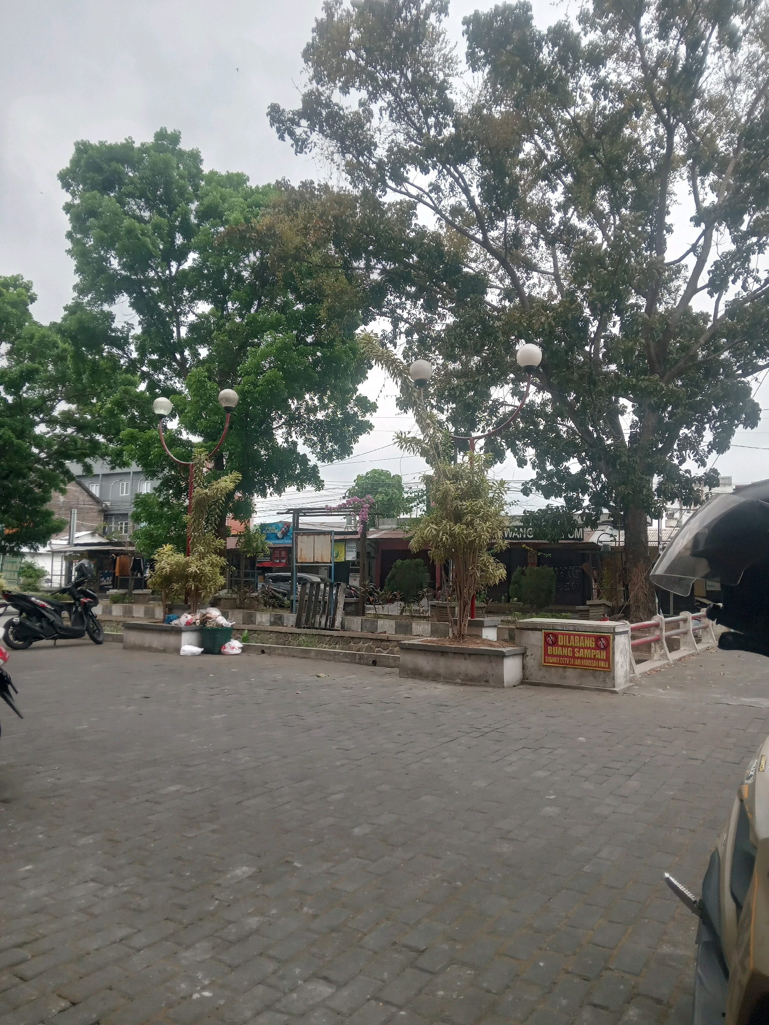 The image captures an outdoor scene, likely in Depok, Indonesia, featuring a paved area with a pedestrian walkway and a street beyond. Dominating the upper half of the frame are large trees with dense foliage, some appearing to be mature. Two white globe-shaped lights on red lamp posts are positioned in the foreground, with a small tree growing between them.
In the middle ground, a collection of shops or stalls line the street, some with signs indicating business activity, one featuring the text "WANG. COM". A trash can filled with plastic bags and debris is visible near the lamp posts. A motorcycle is parked on the left side of the paved area.
A prominent yellow sign with red lettering is mounted on a low barrier in the right-middle ground. The sign reads "DILARANG BUANG SAMPAH" which translates to "NO DUMPING TRASH", and further states "DIAWASI CCTV 24 JAM NONVISUAL AWAD". This suggests a restricted area and surveillance.
The pavement in the foreground is made of interlocking gray bricks. The sky is overcast and appears a uniform light gray. A portion of a motorcycle's reflective windshield is visible in the bottom right corner, indicating the photo was taken from the perspective of someone on a motorcycle. The overall impression is of a somewhat unkempt public space adjacent to commercial establishments.