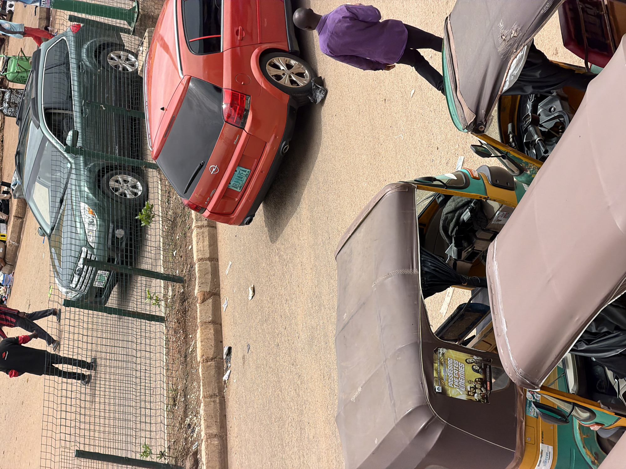 The image depicts a street scene in Bukuru, Nigeria, with several vehicles and individuals present. In the foreground, two auto-rickshaws, or "Keke NAPEPs" as they are known locally, are parked. The one on the left is primarily yellow and green, with a poster attached to its side featuring religious imagery and text. The other auto-rickshaw, partially visible in the foreground, is brown.
Behind the auto-rickshaws, an orange car, identified by its license plate as ABJ-147FA, is parked at an angle. To the left of the orange car, a dark green station wagon is also parked. A man in a purple shirt and black trousers is walking away from the orange car.
Further in the background, near a wire fence, two men are walking. One is wearing a black shirt and black trousers, while the other is in a red and black striped shirt and black trousers. The ground is a mixture of asphalt and sandy dirt. Debris, including paper and plastic, is scattered on the road and sidewalk. The overall impression is one of a busy urban environment with common modes of transportation.