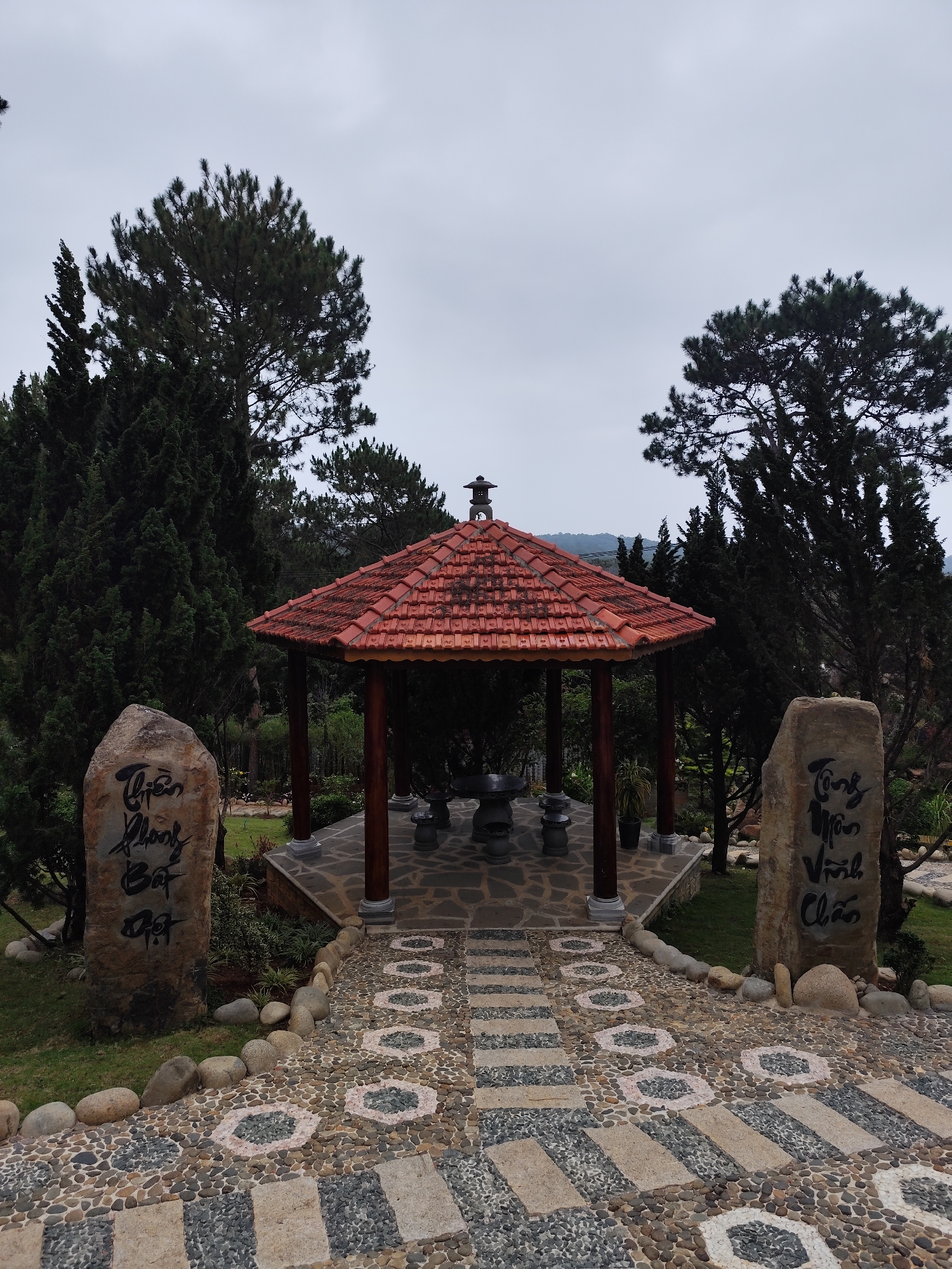 The image depicts a scenic view within a park setting, characterized by a traditional Vietnamese gazebo or pavilion. The pavilion is octagonal with a red tiled roof, dark wooden columns, and a stone platform. Inside the pavilion are stone stools and a table. The area is surrounded by trees, including what appear to be pine trees, with an overcast sky overhead.
A pathway leads to the pavilion, constructed with a pattern of alternating stone pavers and pebble-covered sections. Two large stones with calligraphic inscriptions flank the path, adding a cultural element to the scene. The location is in the city of Da Lat, Vietnam.