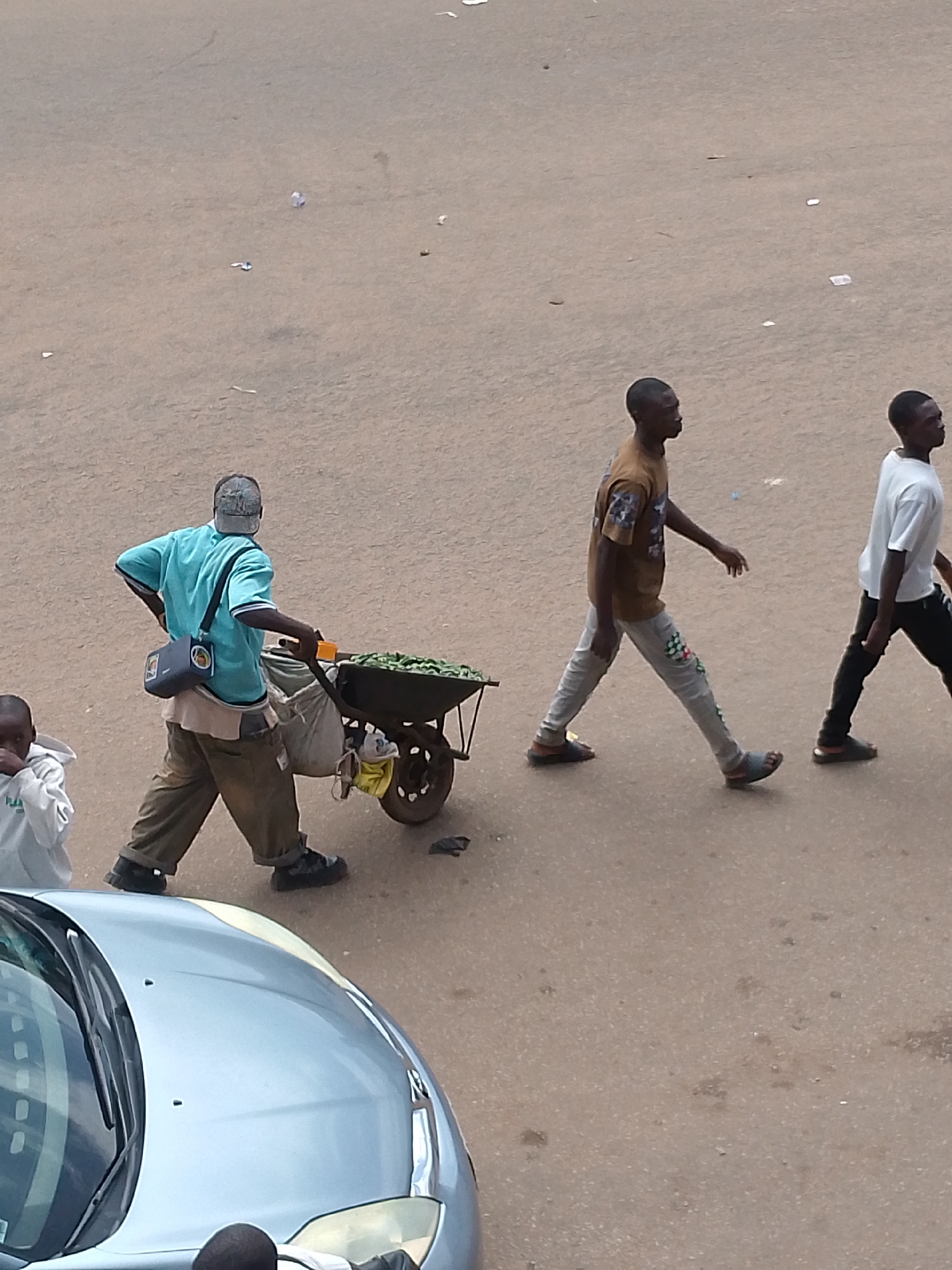 This image depicts a street scene in Bukuru, Nigeria, with four individuals and a car. In the foreground, the hood and windshield of a light blue car are visible, taking up the lower left quadrant. To the left of the car, a child in a white hoodie stands looking away from the camera. Further back, a man wearing a turquoise t-shirt, khaki pants, and a baseball cap is pushing a wheelbarrow laden with green produce. He is also wearing a dark blue bag slung across his chest. To his right, two men are walking away from the camera. The first man wears a brown t-shirt and patterned grey pants, while the second man wears a white t-shirt and black pants. The ground is paved with a gritty, light brown asphalt. Debris, including small white scraps of paper and other unidentifiable detritus, is scattered across the surface. The overall lighting suggests it is daytime.