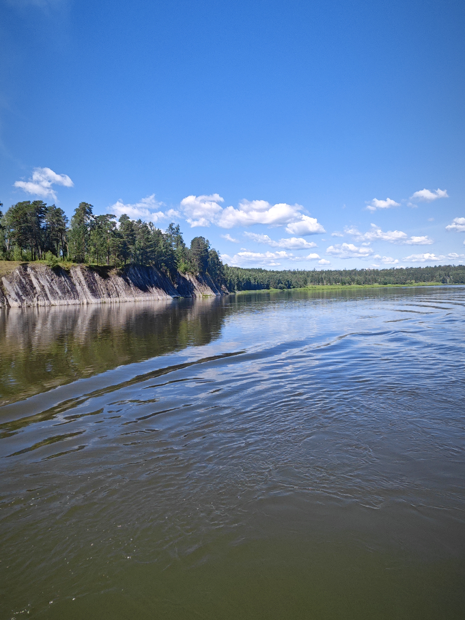 The image shows a wide river on a sunny day. The water is dark green in the foreground and reflects the blue sky and white clouds. There are ripples on the surface of the water, indicating movement. On the left side of the river, there is a steep, rocky cliff covered with trees. The cliff has layered rock formations. The background shows a dense forest stretching across the horizon. The sky is bright blue with scattered white clouds. The location is identified as the city of Savino, Russia.