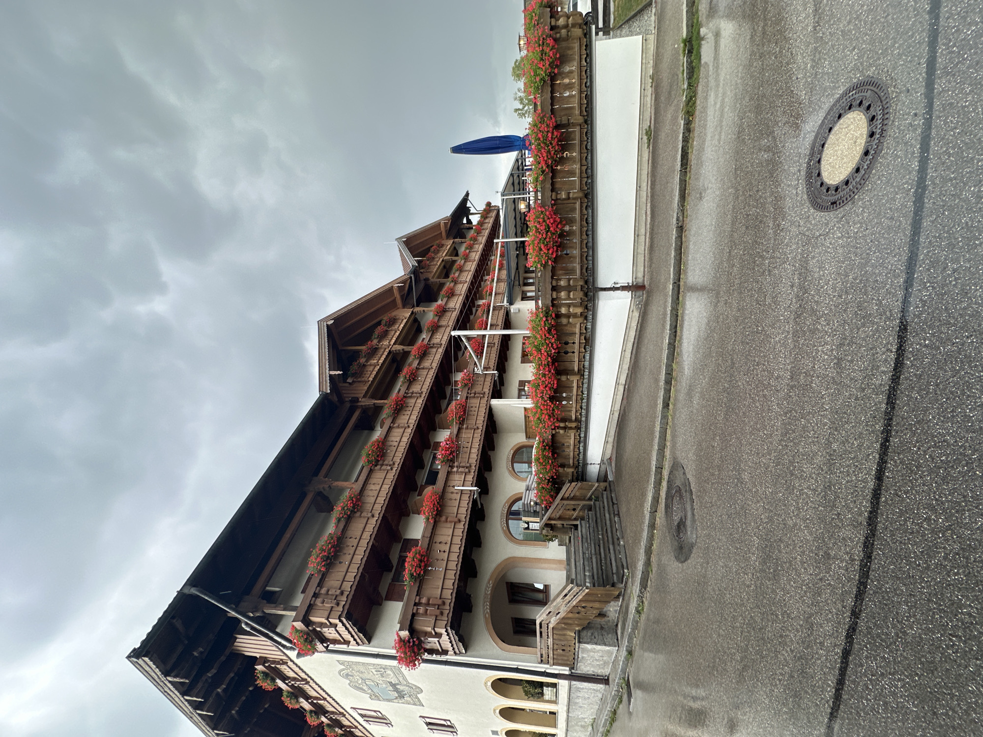 A multi-story building with traditional wooden balconies adorned with red flowers is visible under a cloudy sky. The building, located in Seeon-Seebruck, Germany, features arched entrances on the ground floor and a large set of outdoor stairs leading to the upper levels. The street in front of the building is wet and reflects the overcast sky, suggesting recent rain. A blue umbrella stands near the building's exterior.