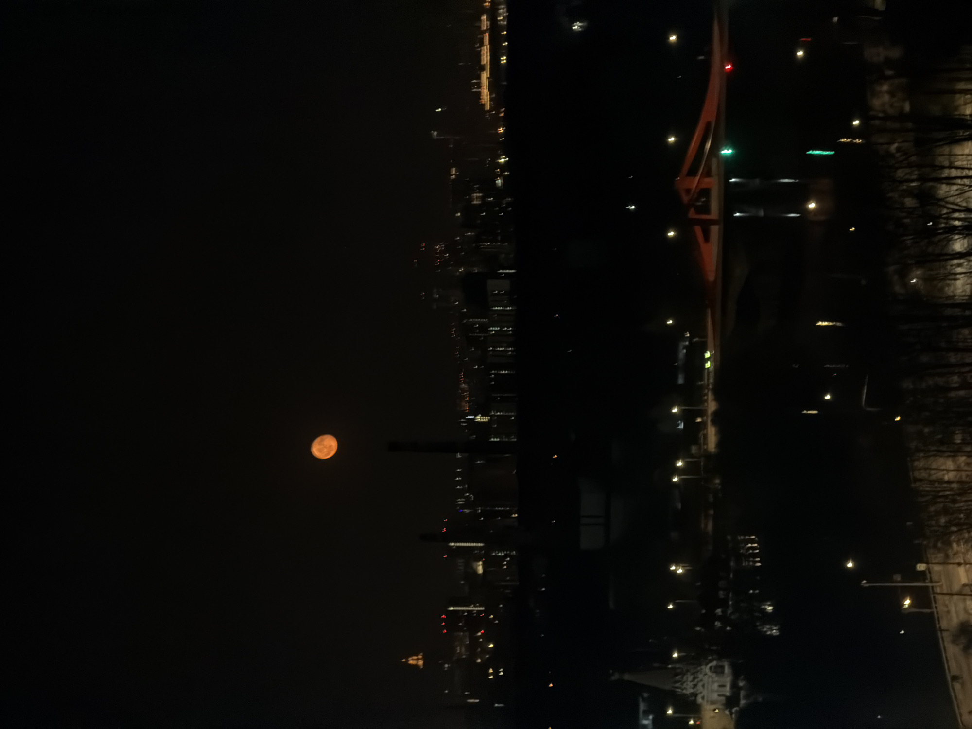 A night view in Khimki, Russia, captures a cityscape illuminated with lights, dominated by a bright orange moon. A red bridge stands to the right, with a darker foreground suggesting nearby trees or foliage. The city lights stretch into the distance, highlighting buildings and structures.