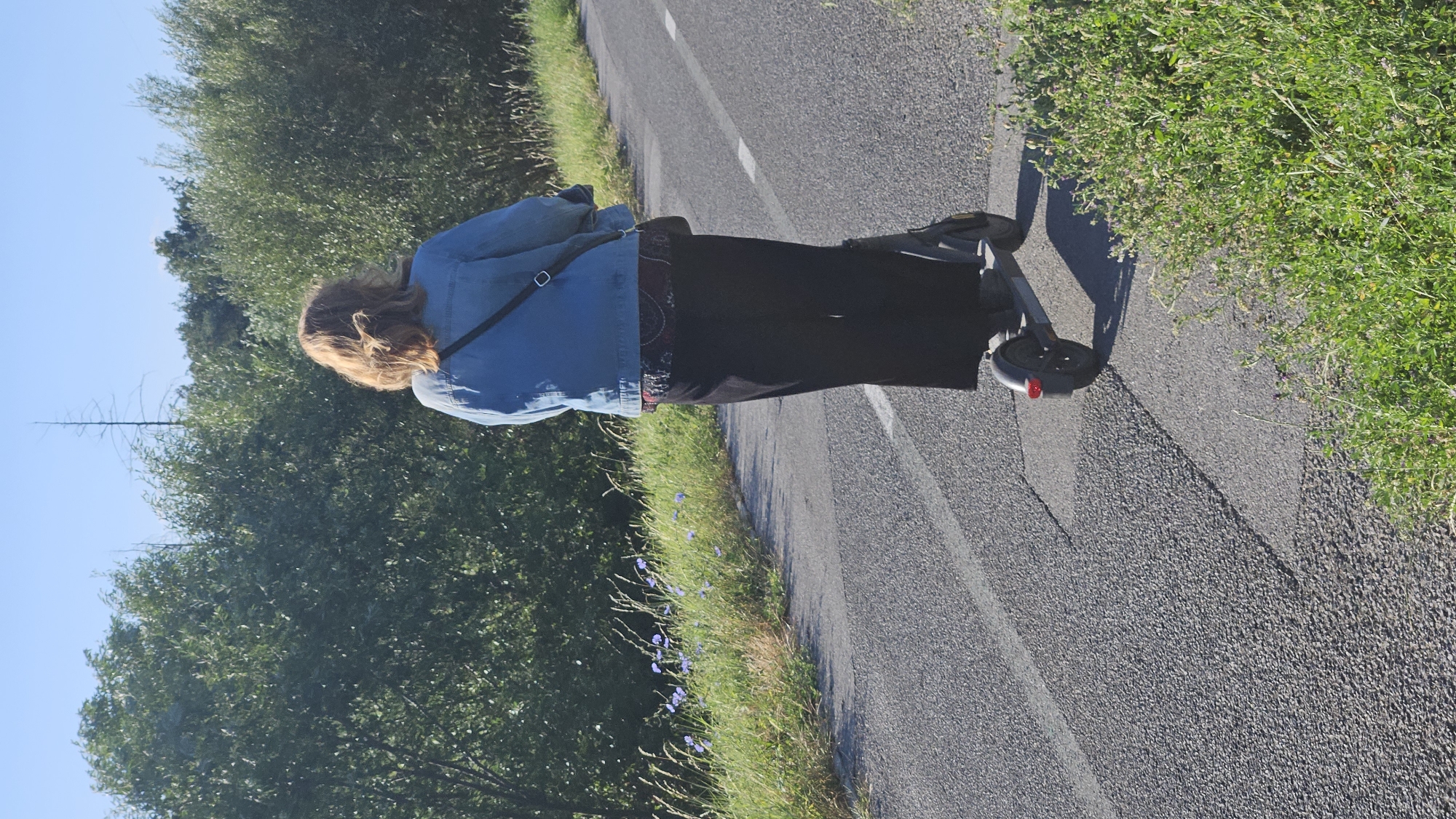 A woman is riding an electric scooter on a paved path. She is wearing a blue denim jacket, a dark patterned top, and black pants. She has her hair tied back. The path is bordered by green grass and trees. The sky is clear and blue. The image appears to have been taken in the city of Prešov, Slovakia.