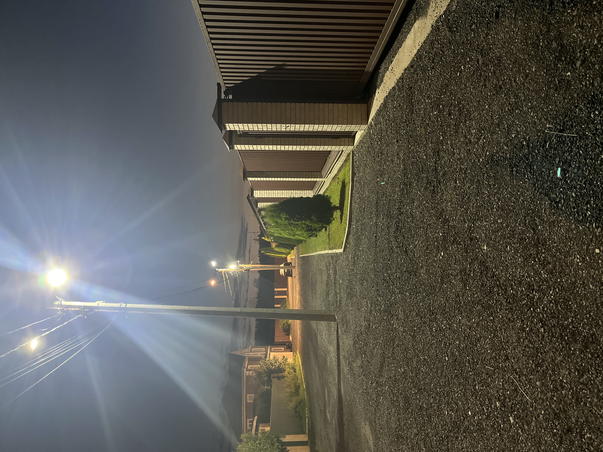 The image shows a street scene at night, likely in a suburban area of Sankt-Peterburg, Russia. A streetlamp illuminates a gravel road that curves to the left. On the right, a tall, dark brown metal fence with brick pillars runs along the side of the road. A line of manicured green bushes borders the fence, followed by a patch of neatly mowed grass. To the left of the road, a green metal fence is visible, behind which is a two-story house with red brick accents. Further down the road, another streetlamp is visible, and a dark-colored car is parked. The sky is dark, indicating nighttime, with some clouds present. The overall impression is that of a quiet, residential street.