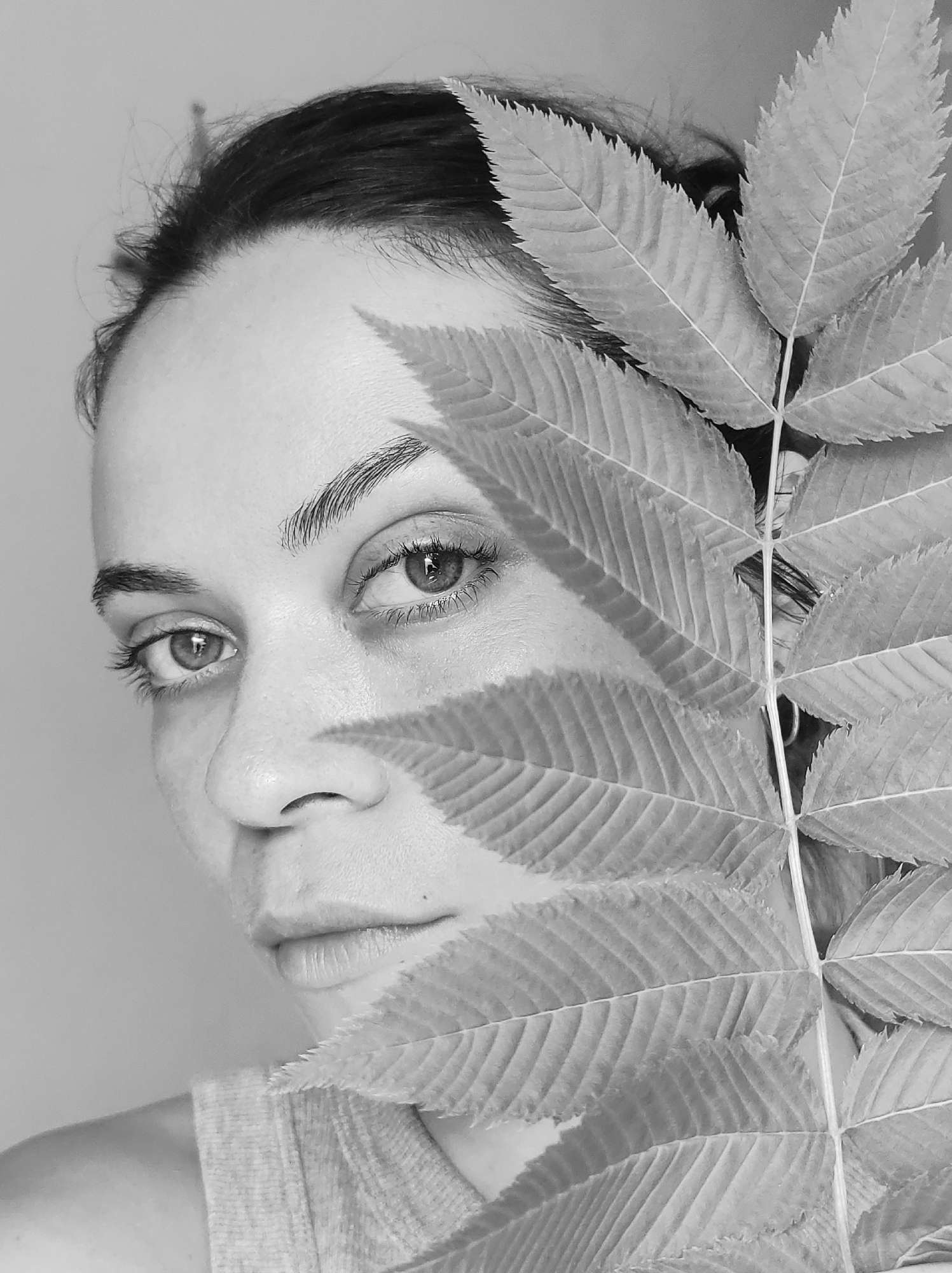 A monochrome close-up portrait features an adult woman, likely in her 20s or 30s, with her face partially obscured by a large, vertically positioned compound leaf. Her dark hair is pulled back, revealing her forehead and hairline. Her left eye (viewer's right) is fully visible, looking directly at the camera with a neutral, contemplative, and slightly intense gaze, reflecting a light source. Her right eye and the right side of her face are largely hidden behind the leaf.
The visible portion of her face shows defined eyebrows, a delicate nose, and full, closed lips. Her skin texture is prominent, suggesting a natural aesthetic. She wears a simple, light-colored top, with a portion of her left shoulder visible at the bottom left of the frame.
The large compound leaf, possibly from a tree like sumac, features a distinct central rachis and numerous oval-shaped leaflets with prominent venation and serrated edges. It is held vertically, covering approximately half of her face.
The lighting is soft and even, highlighting the textures of both the woman's skin and the intricate details of the leaf. The background is a plain, light, undifferentiated tone, suggesting an indoor or studio setting. There are no discernible location cues, time of day, specific weather, or visible text within the image. The overall composition creates an artistic and naturalistic mood.