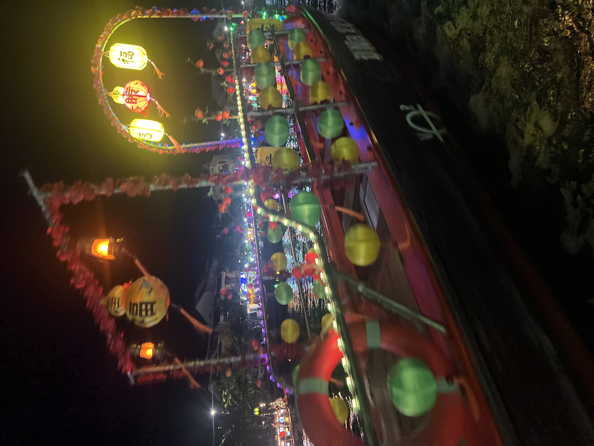This image captures a night scene on the Thu Bon River in Hoi An, Vietnam, specifically during the Lantern Festival. A decorated boat, likely a tourist vessel, is prominently featured, illuminated by numerous colorful lanterns. The boat is adorned with red floral garlands and suspended yellow and green spherical ornaments. A red life preserver is visible on the side of the boat, along with some indistinct Chinese characters painted on its hull. The river itself is dark, reflecting the lights from the boat and the surrounding buildings. In the background, a procession of other boats and the illuminated riverside create a festive atmosphere. The overall impression is one of a vibrant cultural celebration.