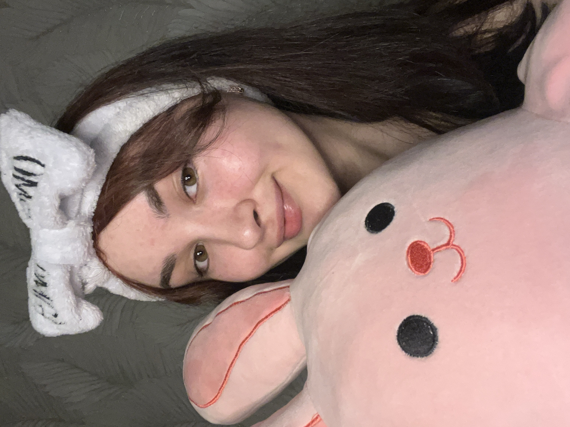 A young woman with brown hair and light skin is lying down, facing the camera. She is wearing a white headband with a bow on top. She is partially obscured by a pink stuffed animal with black eyes and a red nose. The background appears to be a grey textured surface, possibly a bed or couch. No information is available in the image to suggest that it was taken in Neftekamsk, Russia.