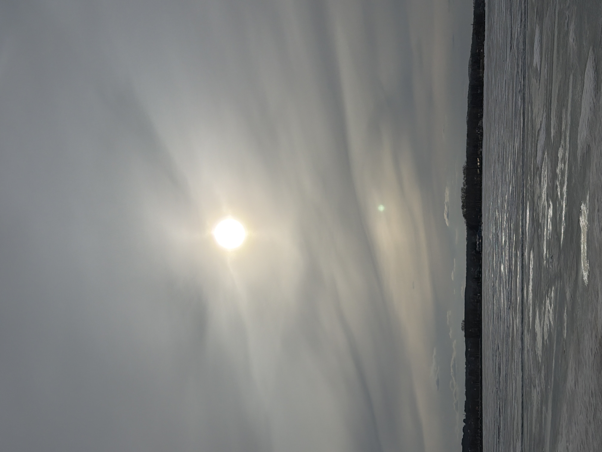 The image depicts a gray, overcast day over a body of water, likely a lake or a wide river. On the right side of the frame is the edge of the water with what appears to be ice partially covering the surface. Beyond the water is a line of trees that suggest the shoreline with buildings on the bank, suggesting a urban area. The sun is visible on the left side of the frame, obscured by clouds and haze, casting a faint light onto the scene. Due to the quality of image, determining the exact location within the city of Votkinsk is challenging. The image does not depict any people. There are no discernible actions or interactions in the scene.