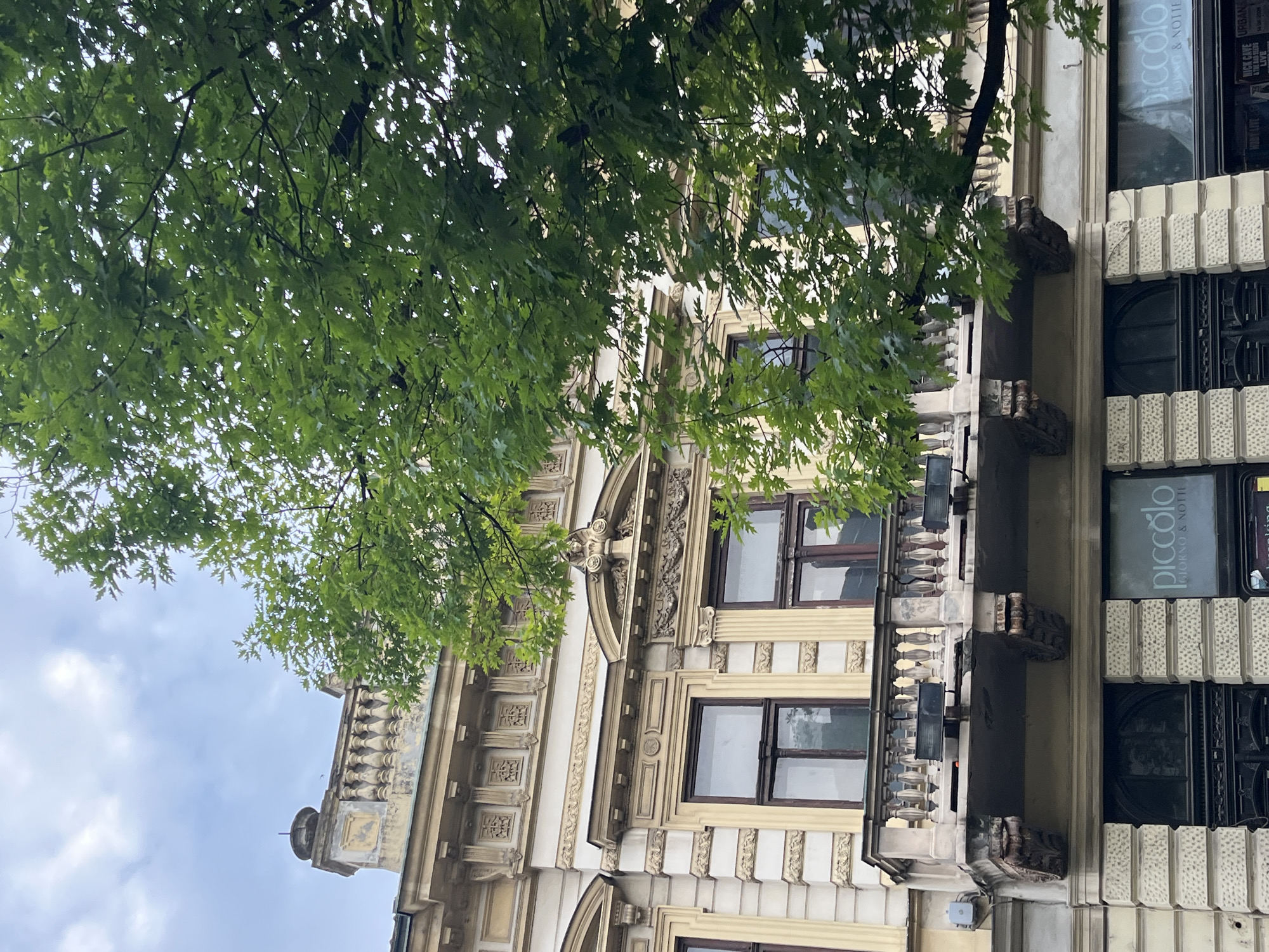 The image presents a detailed view of a multi-story, ornate architectural building in Belgrade, Serbia, partially obscured by a large deciduous tree. The scene is set during the daytime under an overcast or partly cloudy sky.
The building's facade is a light yellow-cream color with white architectural details, showing signs of weathering and age. The uppermost visible section features a stone balustrade atop the roofline, accented by an urn-like finial on a pedestal to the left. Below this, an ornate frieze with repetitive square decorative panels runs along the top story. The windows on the middle floors are framed in dark wood, with some featuring elaborate pediments including classical motifs like swags, scrolls, and sculpted figures. A prominent stone balcony, supported by heavily carved acanthus leaf corbels, extends from the second floor, featuring a balustrade of turned columns.
The ground floor of the building is characterized by a rusticated stone facade. A dark, arched doorway or entrance is visible on the left. To the right, a shop window clearly displays the name "piccolo" in illuminated letters, with "GIORNO & NOTTE" beneath it. Below this, partially visible posters advertise "NICK CAVE THE BAD SEEDS LIVE" and "URBAN".
A large tree with dense, vibrant green foliage occupies the right half of the image, with its branches and leaves extending over the building, partially obscuring the upper and middle sections. The sky above is a pale blue-grey, indicating an overcast or hazy condition. There are no people visible in the image, and no specific actions are depicted. The architectural style and the presence of the "piccolo" restaurant confirm the location as Belgrade, Serbia, likely on Obilićev Venac.