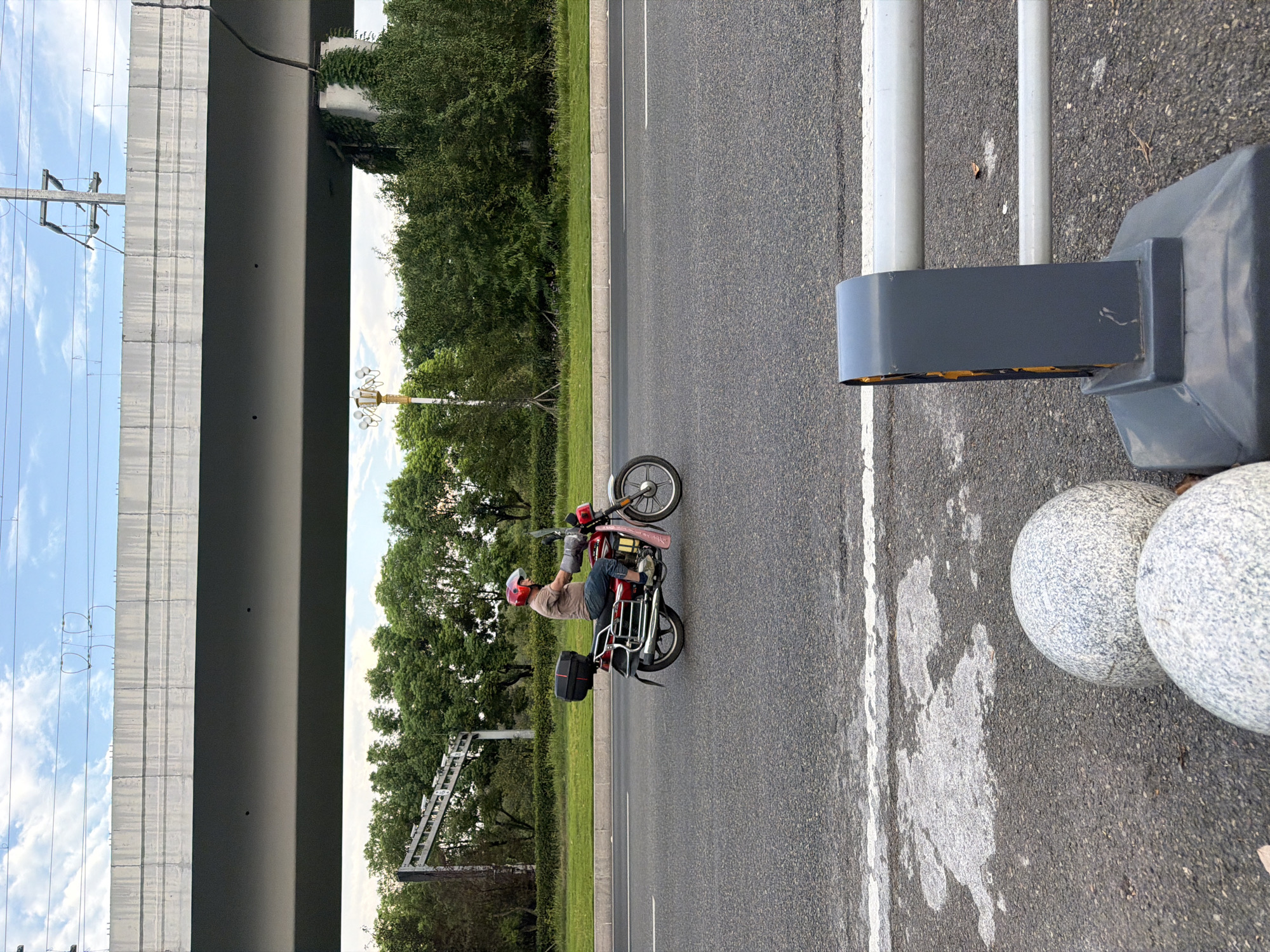 In Jiangjin, China, under an elevated highway overpass, a person wearing a red helmet rides a red motorcycle on a paved road. The rider is wearing a light brown short-sleeved shirt, blue jeans, and gloves. The motorcycle has a black storage box on the back and a pink fender. Trees and greenery line the side of the road, and overhead power lines are visible. In the foreground, a grey bollard with a white bar is situated on the road.