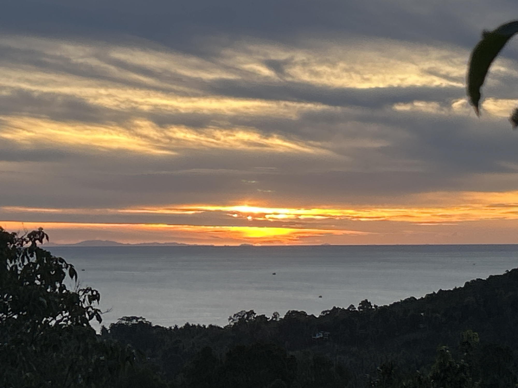 The image depicts a sunset over the ocean. The sun is partially obscured by clouds, creating a radiant glow. In the background, an island is visible on the horizon. The foreground features lush green trees and foliage. The location is Baan Sri Thanu, Thailand, as evidenced by the typical vegetation and coastal landscape. There is no indication of people.