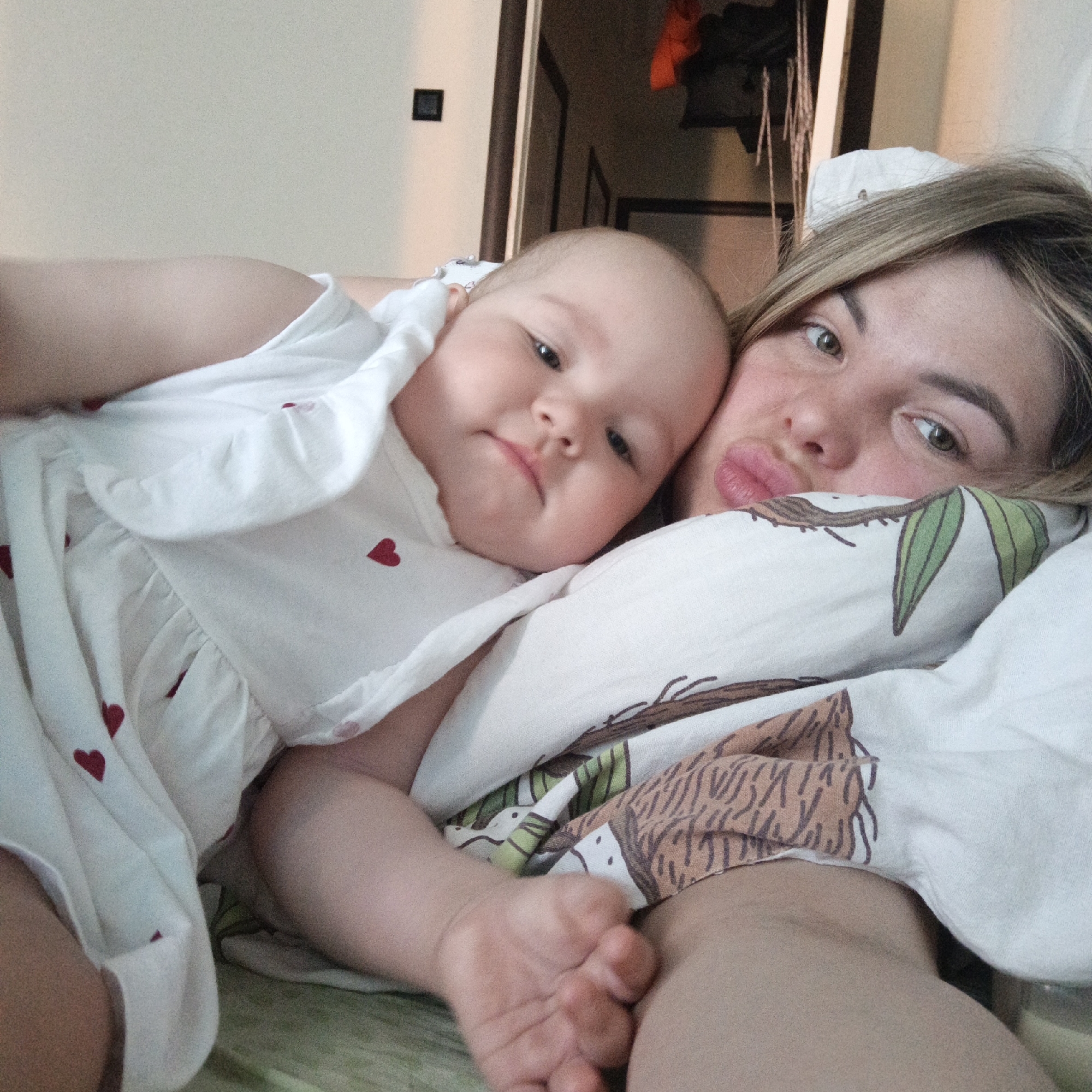 The image shows a light-skinned woman and a baby lying in bed. The woman is on her back, with her head on a pillow covered in a brown and green leaf pattern. She has light blonde hair and is looking at the camera with slightly pursed lips. The baby, wearing a white dress with red heart accents, is lying on her chest. The baby is looking at the camera with a slightly furrowed brow. The bedding is a light green color. In the background, there is a doorway and a wall with a black electrical outlet. The location is Unknown, Unknown.