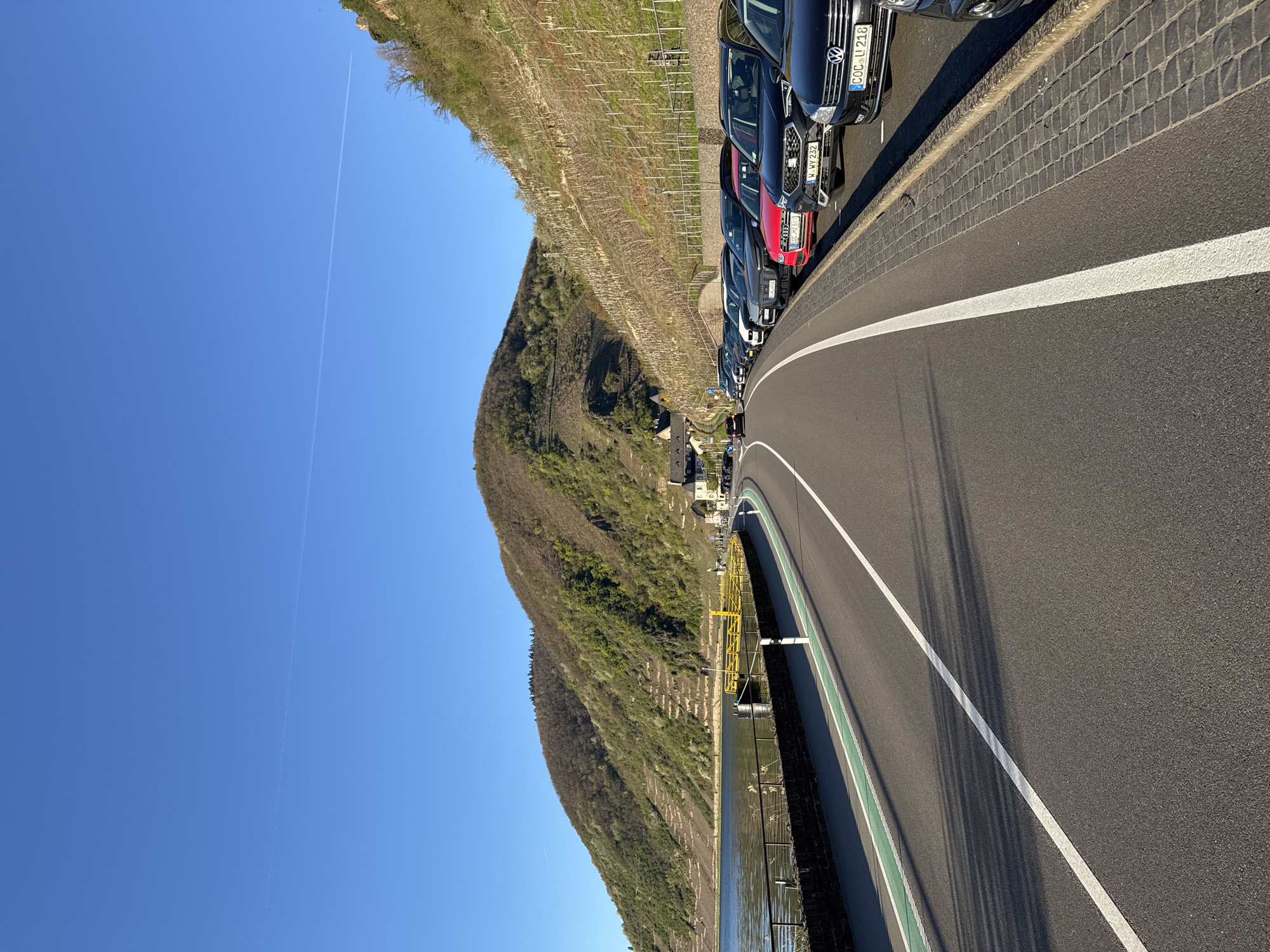 The image shows a road with parked cars along the edge in Beilstein, Germany. A mountain covered with green vegetation is seen on the left. A line of cars, including a Volkswagen and an Audi with license plates beginning with "COC" and "W," respectively, are parked along the right edge of the road. White lane markings are present on the road. A river with a protective barrier is visible next to the road. The sky is blue with a contrail. The area adjacent to the mountain appears to be a vineyard. A small settlement of buildings is visible midway between the mountainside and the parked cars.