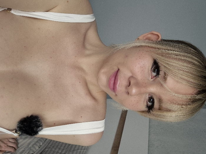 The image displays a close-up, chest-up portrait of a female individual situated indoors, specifically in what is stated as the city of Batumi, Georgia. The subject has fair skin, marked with numerous visible freckles on her nose and cheeks. Her hair is light blonde, styled with bangs covering her forehead and textured, shorter hair on the sides. Her eyes are brown, accented with dark eyeliner and mascara. Her lips are full and colored with pink lipstick, exhibiting a faint, closed-mouth smile while she gazes directly at the camera. A small, light-colored stud earring is present in her right earlobe. She is wearing a white garment with visible shoulder straps. A small, spherical, black fluffy object is clipped to the right strap of this garment. The immediate background features a plain light grey wall. To the right, a segment of a light-colored headboard and striped fabric, consistent with bedding, are visible, indicating a bedroom environment.