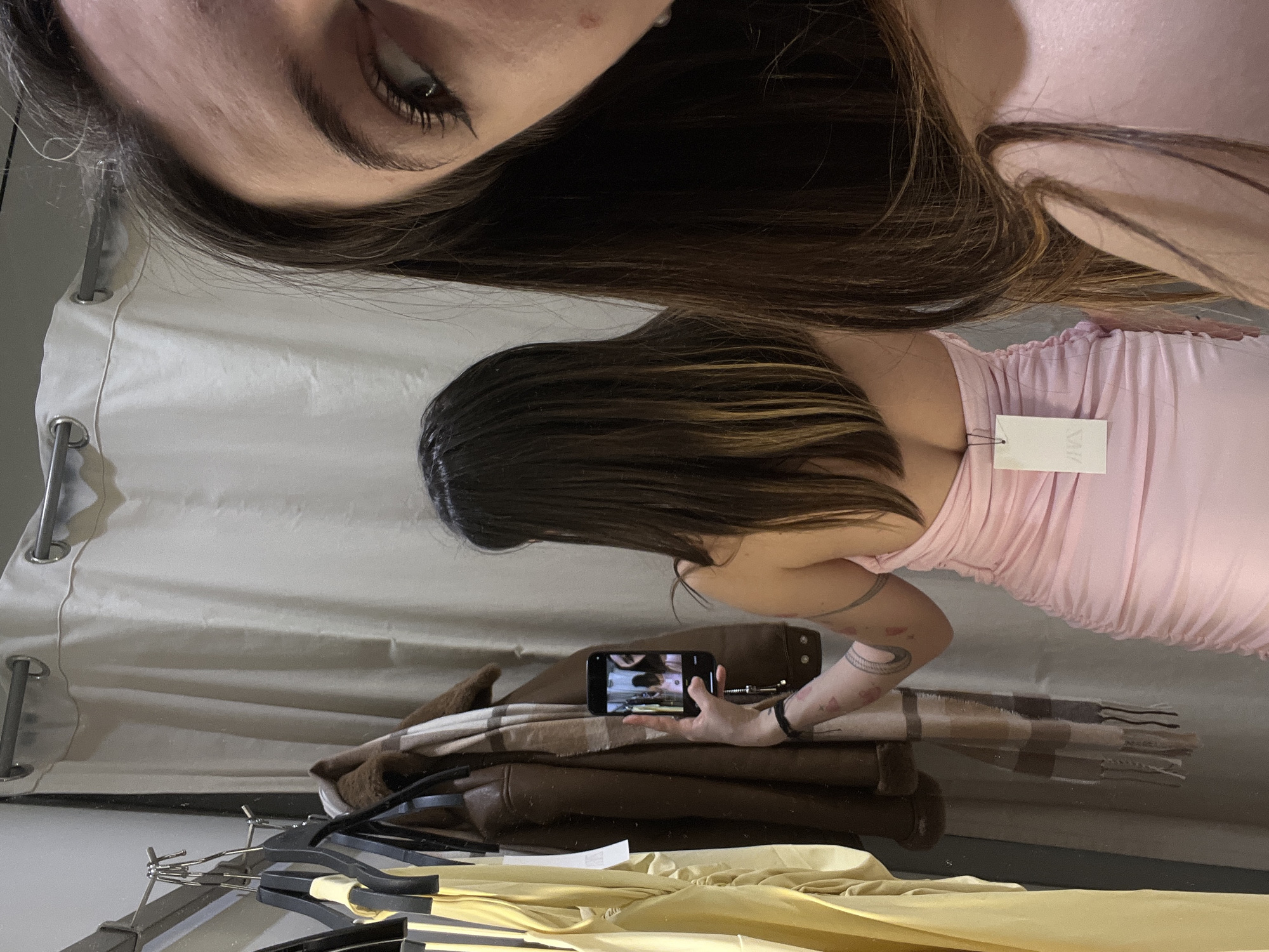 The scene depicts two individuals in a fitting room, likely within a Zara retail store in Riga, Latvia. One individual is seen from behind, wearing a light pink, strapless, ruched dress with a "ZARA" brand tag prominently displayed on the upper back. This person has long, dark brown hair with lighter highlights towards the ends and a tattooed left arm, featuring a crescent moon and abstract designs. A second individual is partially visible in reflection on the right, showing the right side of their face, including an eye and forehead, with long dark hair. This second individual holds a black smartphone in their left hand, pointed towards the first person, capturing an image or video; the phone screen itself displays a reflection of the two subjects. To the left, a metal clothing rack holds several garments, including a yellow item, a brown jacket with faux fur lining, and a plaid scarf. A light beige fitting room curtain hangs in the background.