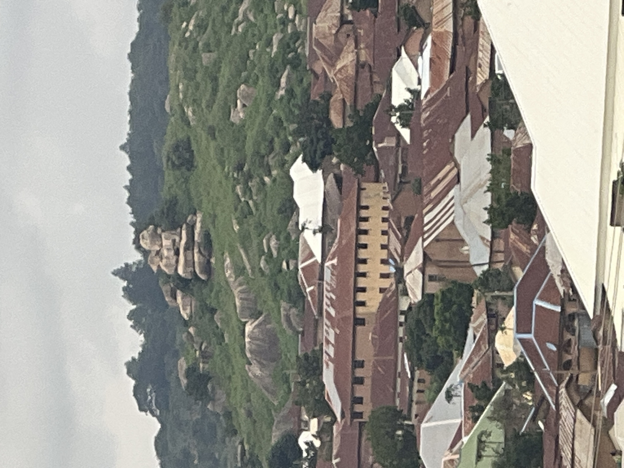 The image shows a cityscape in Jos, Nigeria, dominated by a rocky, green hill in the background. The hill features prominent, weathered rock formations. In the foreground, a dense collection of buildings with predominantly red, corrugated iron roofs covers the landscape. Several buildings are visible with multi-story structures and numerous windows. Some white-roofed structures are also interspersed among the red-roofed buildings. Trees are scattered throughout the urban area, with denser vegetation on the hill. The sky is overcast and appears cloudy. The scene depicts a densely populated urban environment nestled against a natural hilly terrain.