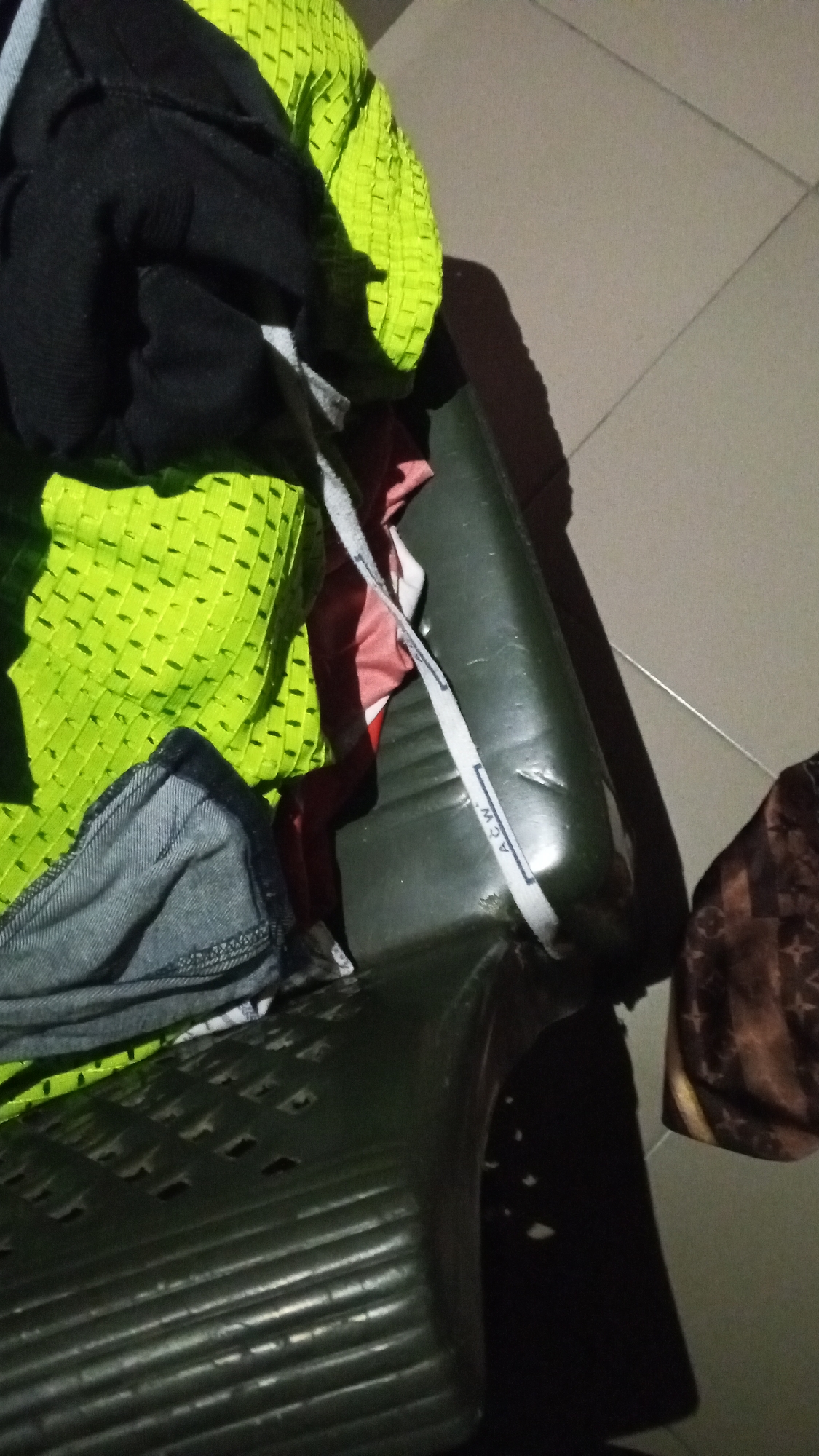 The image shows a close-up view of a dark green, ribbed plastic chair with several pieces of clothing draped over it. The clothing includes a bright neon green mesh-like fabric, a black garment, a section of denim fabric, and a piece of red and white patterned fabric. A white strap with the text "ACW" is visible across the seat of the chair. The chair appears to be indoors, with a tiled floor visible in the background. A shadow is cast on the floor to the right of the chair. The lighting is dim, suggesting it might be evening or an indoor setting with low light. There are no people visible in the image. The scene is consistent with a domestic setting in Jalingo, Nigeria, where such chairs are commonly found and laundry is often placed in open areas or on furniture.