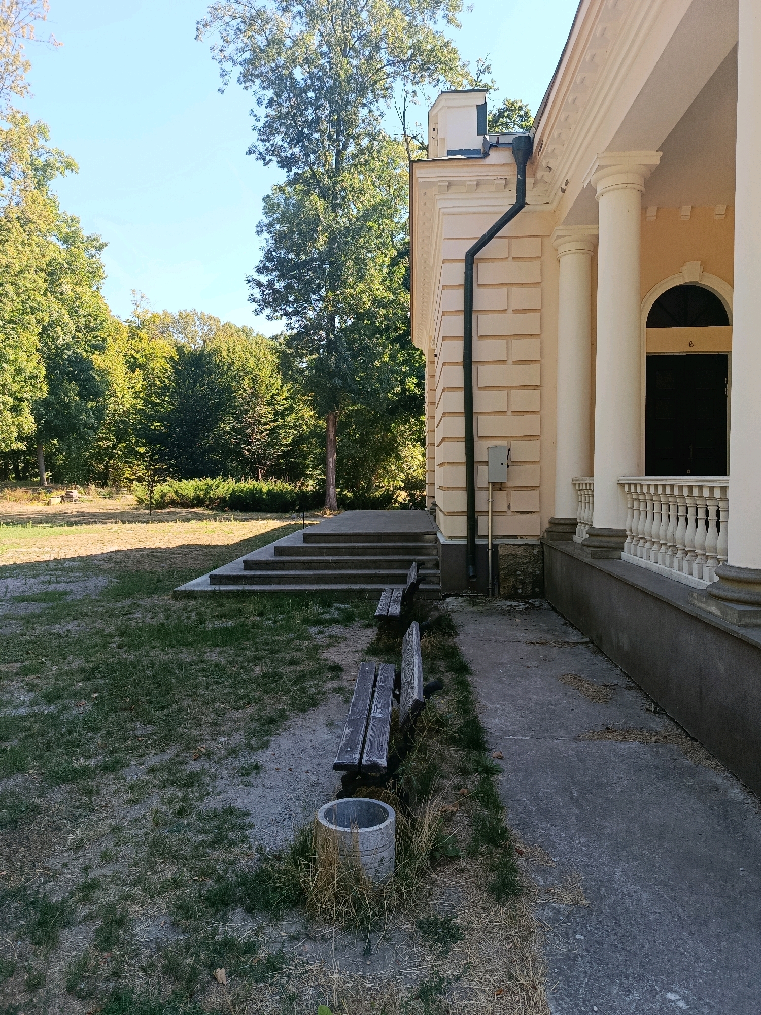 The image depicts the side view of a pale yellow building with white columns and ornate detailing, likely a historical or public structure. The building has a dark arched doorway and a white balustrade along its facade. A dark downspout runs vertically along the textured wall of the building. In the foreground, a concrete pathway runs parallel to the building. To the left of the pathway are two weathered wooden benches placed on a patchy lawn with dry grass and scattered leaves. Adjacent to the benches is a circular concrete structure, possibly a planter or a base for a bin. Beyond the immediate foreground, a set of concrete steps leads up to the building. The area around the building is surrounded by mature trees with green foliage, suggesting a park-like setting. The sky is clear and blue. The location is identified as Bila Tserkva, Ukraine.