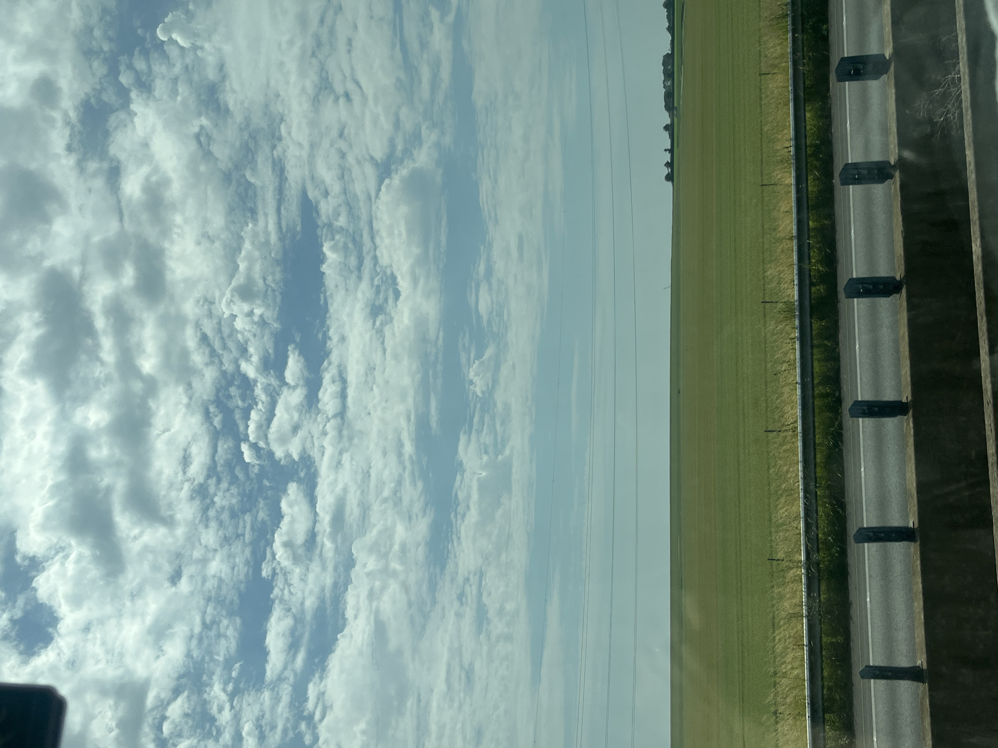 An expansive view of a green field under a cloudy sky is captured from a roadside perspective. The field, likely agricultural, stretches horizontally across the frame, meeting a hazy horizon. In the distance, faint structures suggestive of a rural settlement are visible. A grey highway barrier with vertical bollards in dark grey runs along the foreground, separating the viewer from a paved road. The sky is filled with dynamic white and grey clouds, with patches of blue peeking through. The image appears to be taken from a moving vehicle, as suggested by the blur of motion in the foreground elements. The location is identified as Cuvillers, France.