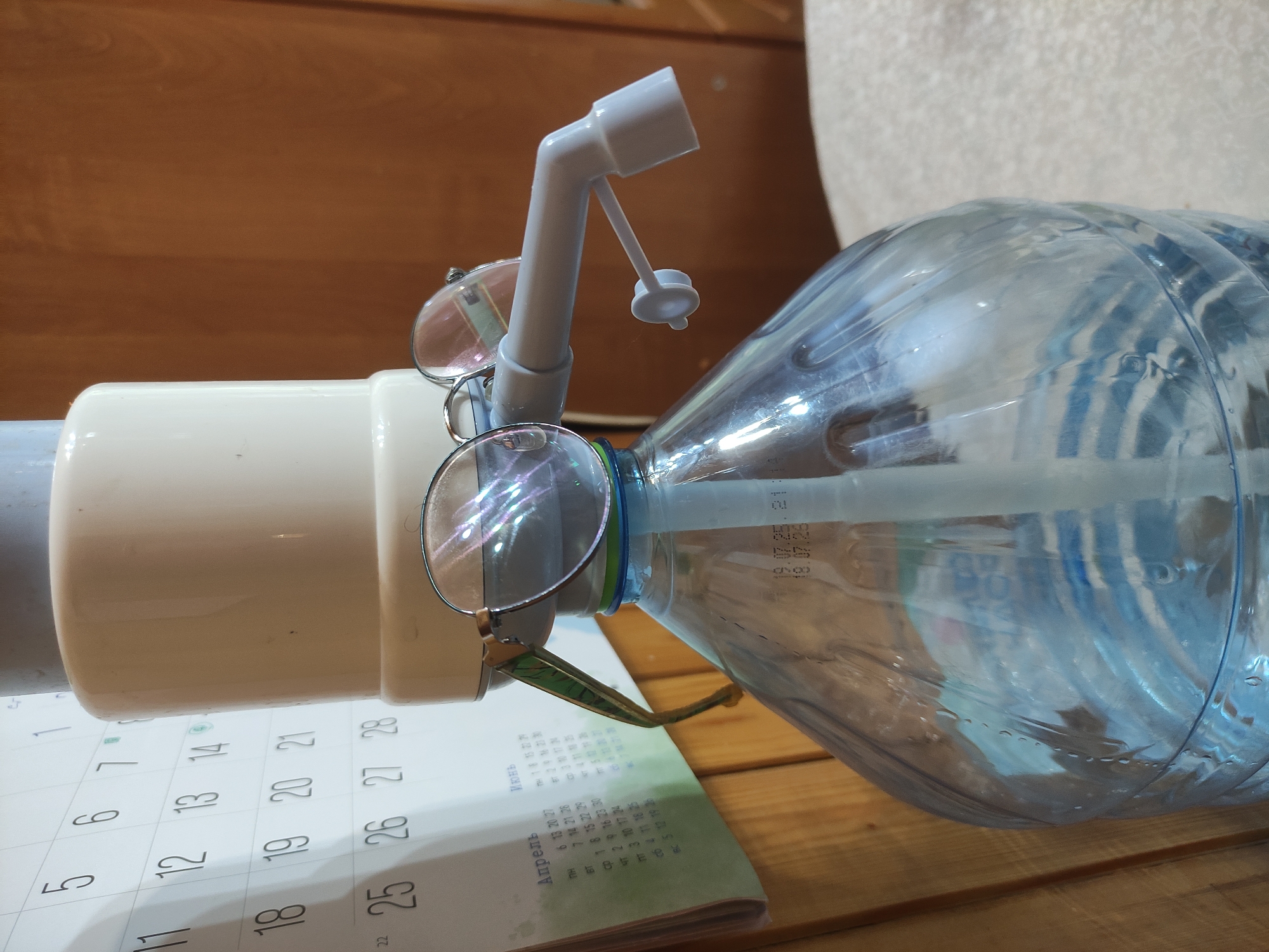 A photograph shows a plastic water bottle with a white plastic pump attached, placed on a calendar that is laying on a wooden surface. A pair of eyeglasses is perched on the pump and the bottle's neck. The calendar shows dates from April and June, and the numbers 11 through 28 are visible. The bottle contains water and is partially obscured by the glasses and the pump. A date stamp is visible on the bottle: "19.07.25.21:14 18.07.26". The background includes a wooden panel and a glimpse of a white surface, possibly a wall or furniture. The photo appears to be taken indoors. There is no information in the image to establish a location in Kratovo, Russia.