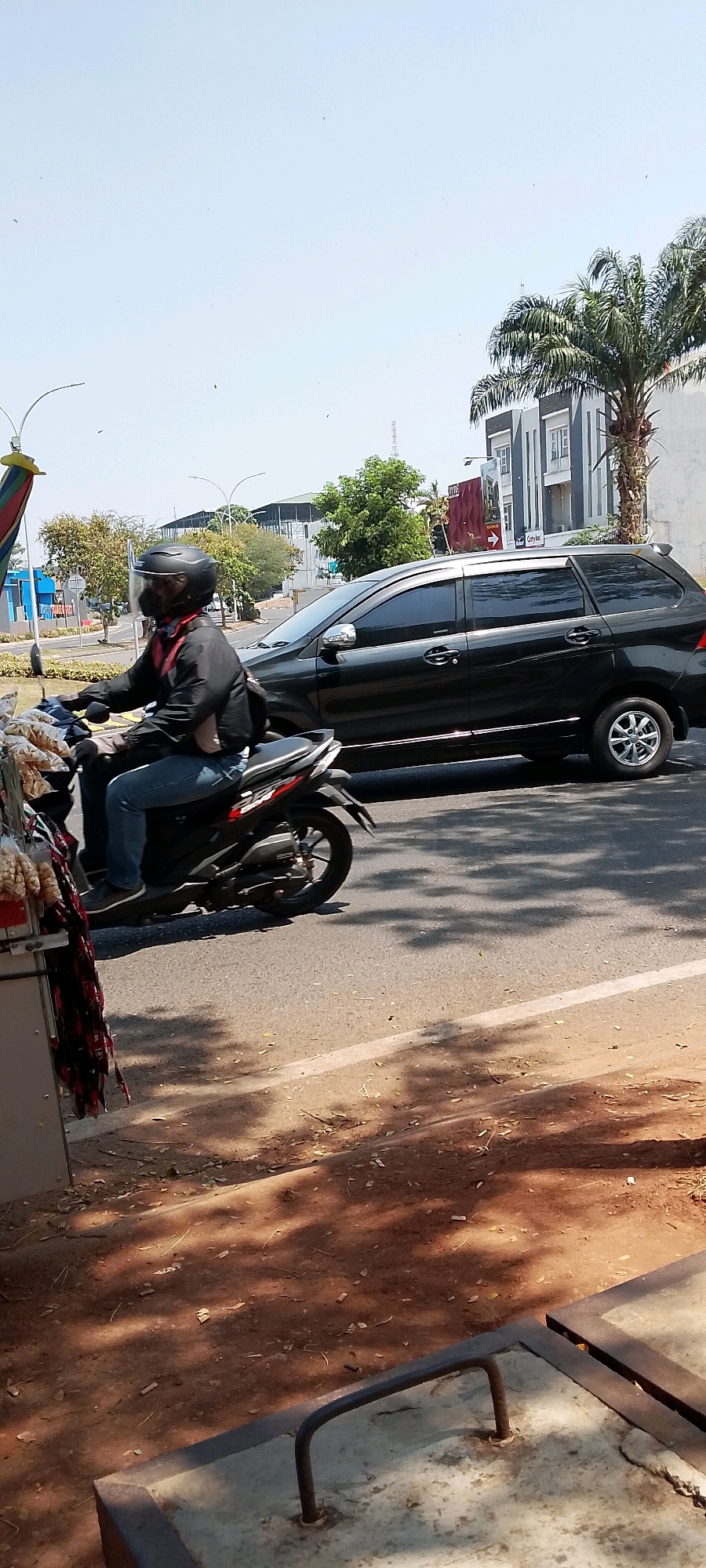 The image depicts a street scene in Gunung Putri, Indonesia, on a sunny day. In the foreground, a vendor's cart is partially visible on the left, displaying various items including what appears to be snacks. A person wearing a black helmet and a black jacket over a red garment is riding a black motorcycle. The person is wearing blue jeans and dark shoes. The motorcycle is moving along an asphalt road. To the right of the motorcycle, a black car is also moving on the road. In the background, there are buildings, trees, including a palm tree, and a clear blue sky. The road appears to have a white line marking. Shadows of trees are cast on the road and the ground in the foreground. The ground in the foreground is reddish-brown earth covered with fallen leaves.