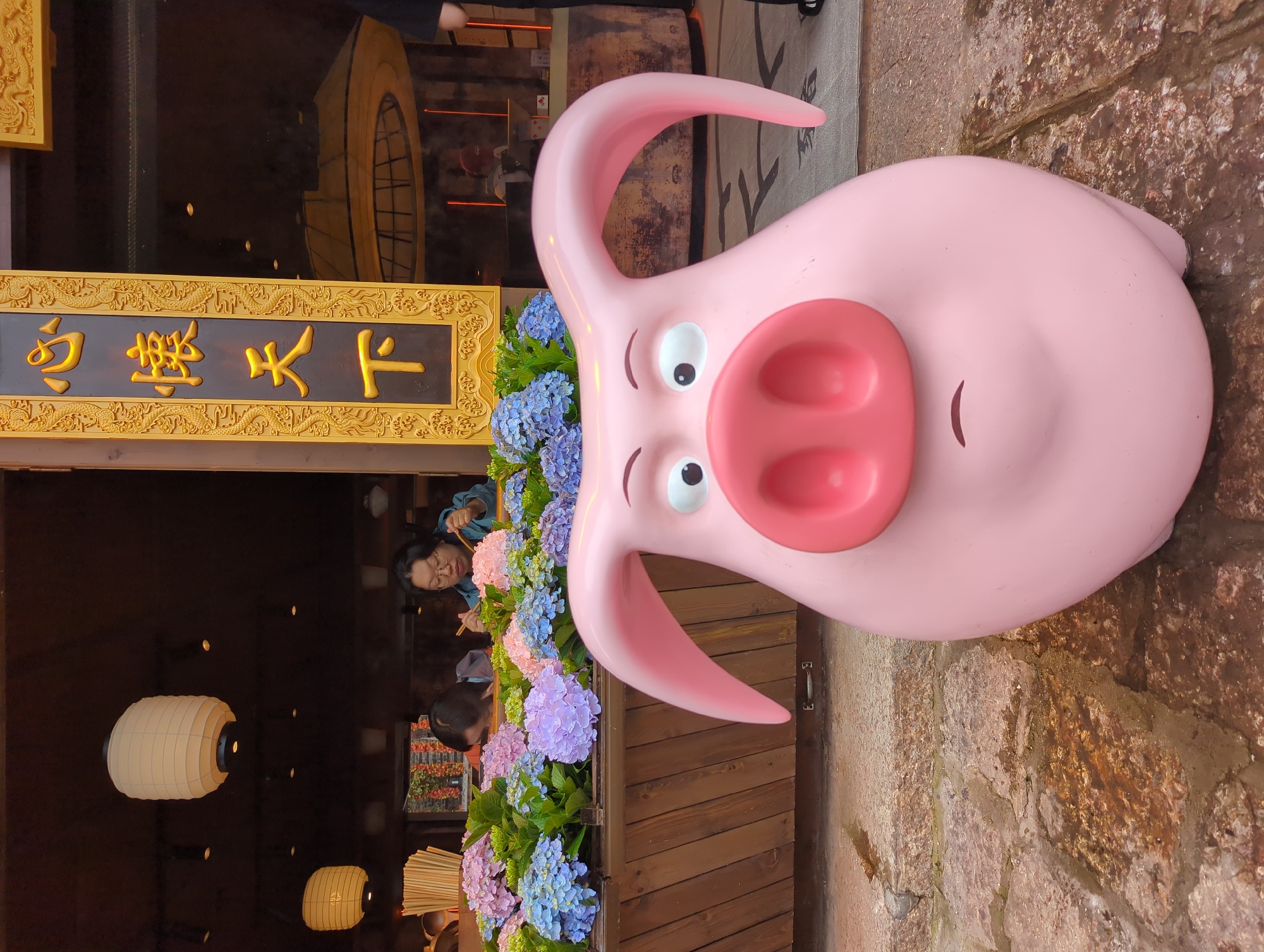 A pink pig sculpture with wide eyes, a large snout, and downturned ears is positioned in the foreground on a cobblestone path. Behind the sculpture, a wooden planter filled with vibrant pink, blue, and purple hydrangeas is visible. In the background, two women are seated at tables, one eating with chopsticks, and a vertical sign with gold Chinese characters is mounted on an ornate golden frame. The setting appears to be a restaurant or shop in Gucheng District, China, with traditional lanterns hanging indoors.