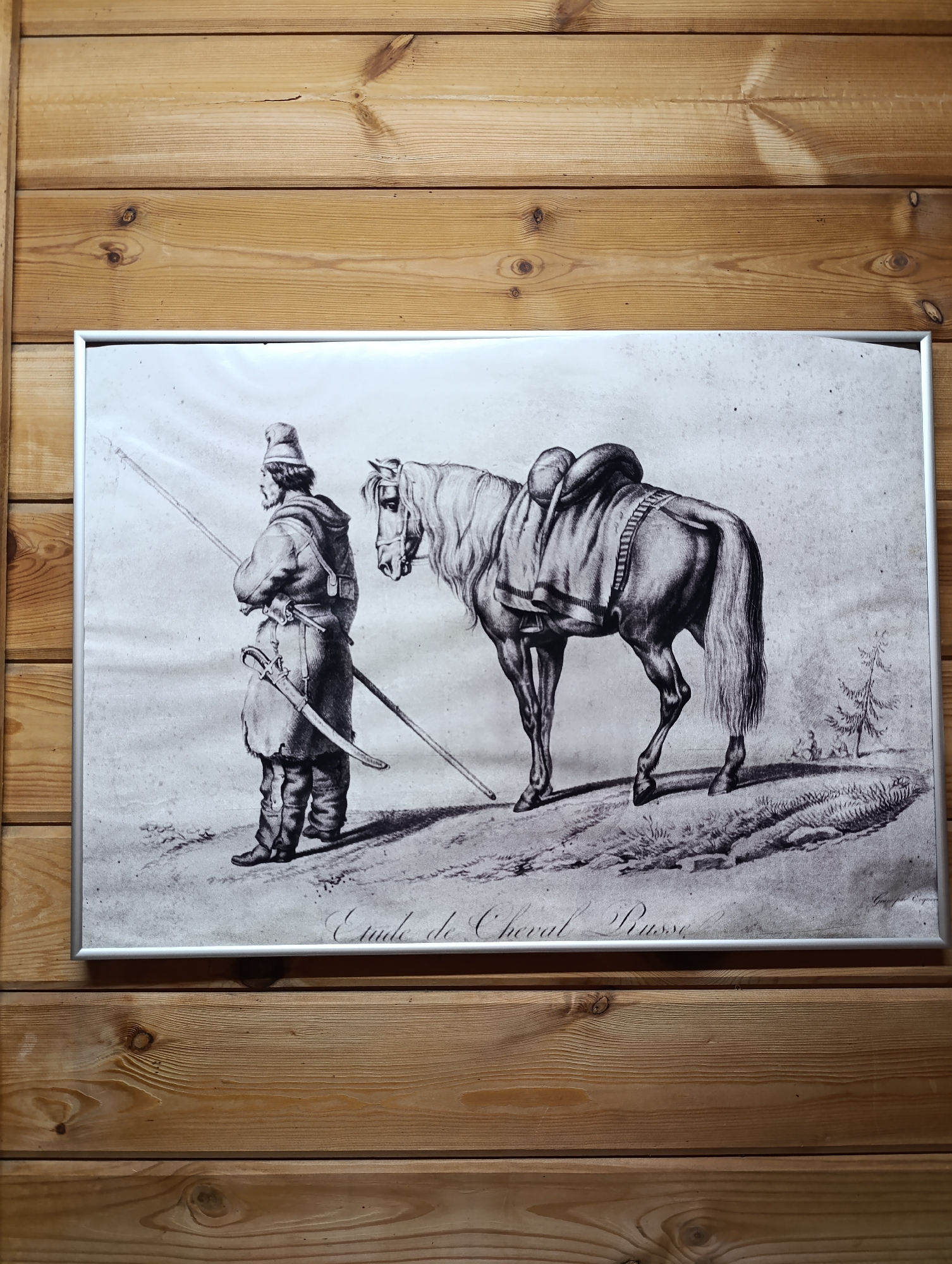 A framed black-and-white print hangs on a wooden wall. The print depicts a man in traditional clothing, possibly a Cossack, standing next to a horse. The man holds a long spear and has a saber at his side. The horse is saddled. The print includes the inscription "Etude de Cheval Russe". The wooden wall consists of horizontal planks with visible wood grain and knots. The location is not determinable from the image.