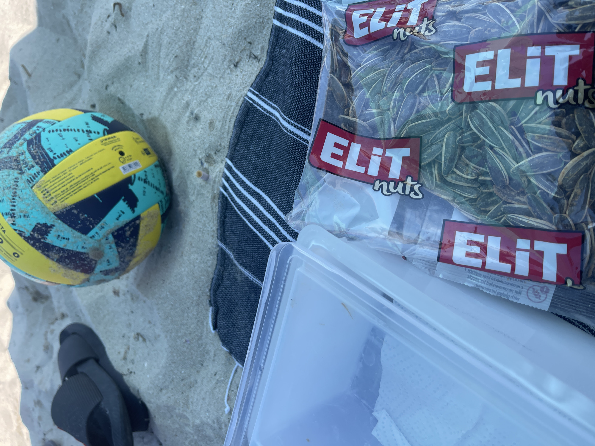 A group of items, including a volleyball, a pair of black flip-flops, a towel, a plastic container with white paper napkins, and a large bag of sunflower seeds, are scattered on the sand at the beach in Kranevo, Bulgaria. The volleyball is yellow, blue, and black. The towel is dark gray with white stripes. The bag of sunflower seeds has the brand name "ELIT" in red and white letters. The scene suggests a beach outing or picnic.