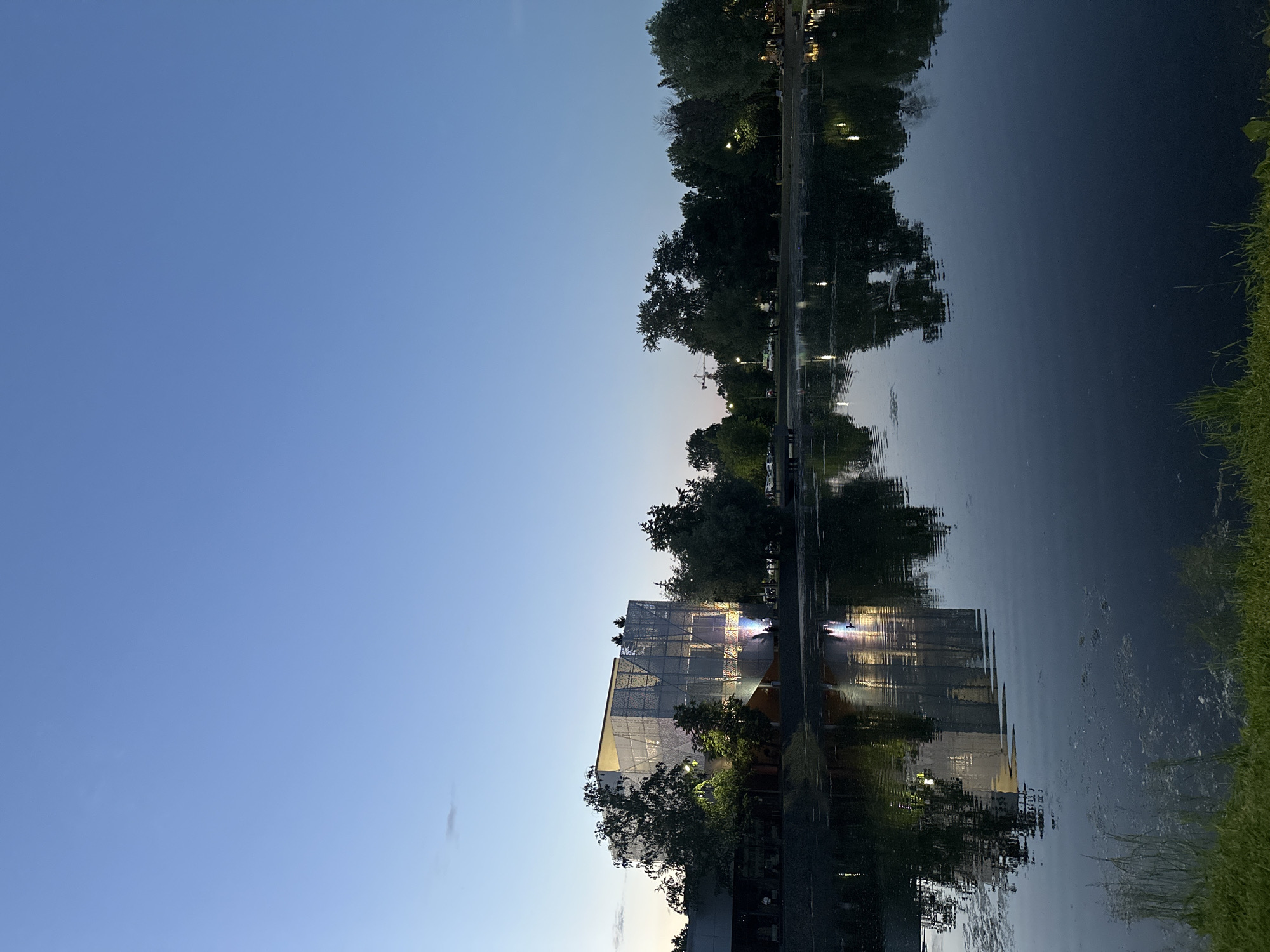 The image, when correctly oriented, presents a serene waterside landscape at dusk or early night in Kazan, Russia. A calm body of water, likely a river or canal, dominates the right half of the scene, perfectly reflecting the evening sky and the structures on the opposite bank.
On the left bank, a distinctive modern building stands out with a unique, possibly perforated or mesh-like, facade. Interior lights illuminate the building, casting a warm glow and creating intricate patterns that are mirrored in the water below. Surrounding the building and lining both banks of the water are numerous deciduous trees, their dark silhouettes contrasting against the twilight sky and their forms reflected with remarkable clarity on the water's surface. Faint artificial lights, possibly streetlights or lights from distant establishments, punctuate the far bank's tree line, adding further luminous reflections to the water. A subtle gradient of blue hues marks the sky, transitioning from a lighter tone near the horizon to a deeper blue overhead. A small patch of green grass is visible along the closest bank. In the distance, above the trees on the far bank, a construction crane is faintly discernible against the horizon. The overall atmosphere is tranquil and contemplative, with no specific activity taking place and no people or discernible text visible.
