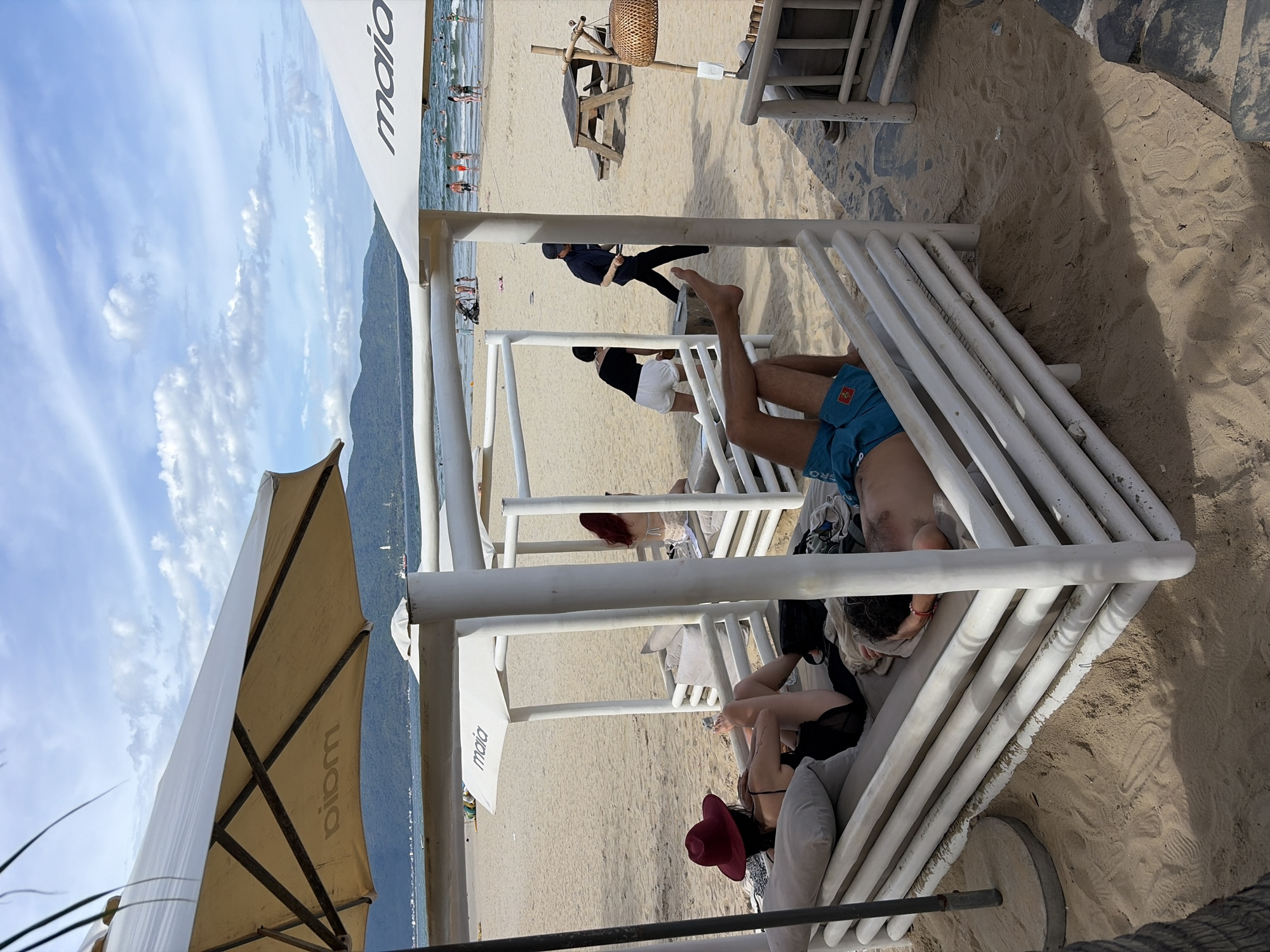 The image depicts a beachfront scene at a resort, likely in the Mỹ Khê Beach area of Đà Nẵng, Vietnam. In the foreground, a white, slatted lounge structure on the sand is occupied by two individuals. One person, seen from behind and wearing a red wide-brimmed hat and black swimwear, is reclining on cushions, holding a drink. Another person, also seen from behind, is lying on their stomach with their head resting on their arms. A third person is lying on their back, facing away from the camera, wearing blue swimming trunks.
In the middle ground, other lounge structures and patrons are visible on the sandy beach. Further back, the sea meets the shore, with people swimming and walking in the water. The background features a hazy mountain range under a partly cloudy sky. Two large white umbrellas, one with the logo "maia" visible, provide shade over sections of the beach. A wooden picnic table and benches are situated on the sand to the right. The overall atmosphere is relaxed and tropical, typical of a beach resort.