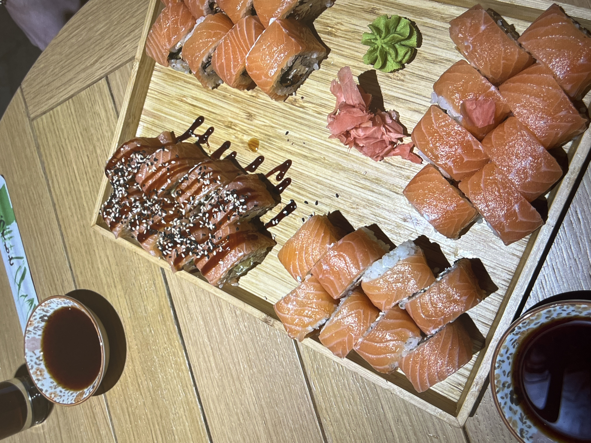 A wooden tray filled with various types of sushi is placed on a wooden table. On the tray, there are several rows of sushi: one row appears to be topped with eel sauce and sesame seeds, another row consists of salmon rolls, and there is also a portion of wasabi and pickled ginger. Two small bowls filled with dark sauce are on the sides of the tray. One set of chopsticks is also partially visible. The location is Unknown.