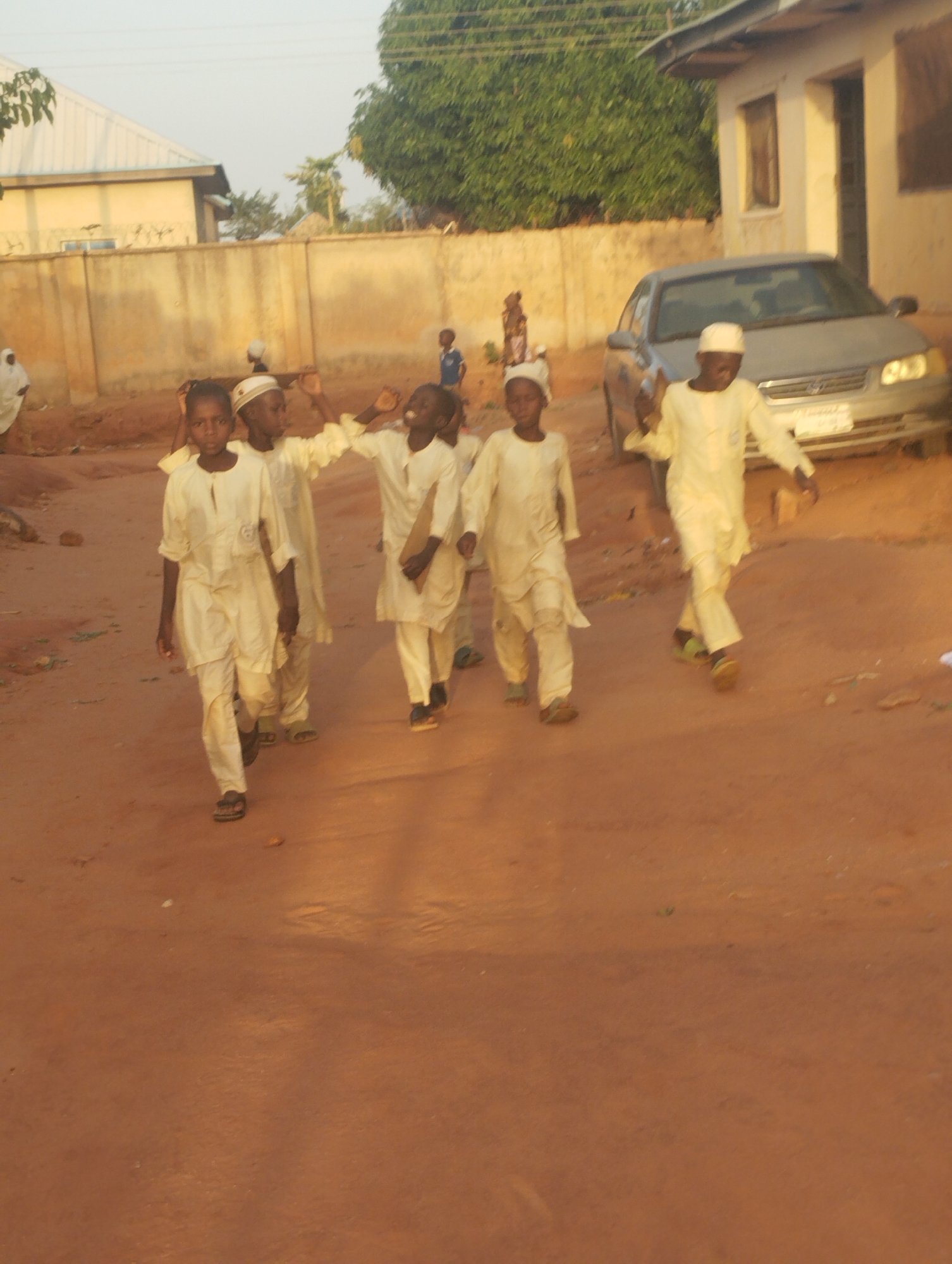 Five children wearing matching white outfits and caps walk along a red dirt road in Tumfure, Nigeria. A silver sedan car is parked on the right side of the road, near a building with a dark doorway. Behind the children, a tan wall stretches across the frame, with another building visible in the background on the left. Other figures stand in the distance. A tree provides foliage in the background, and the overall light suggests either early morning or late afternoon.