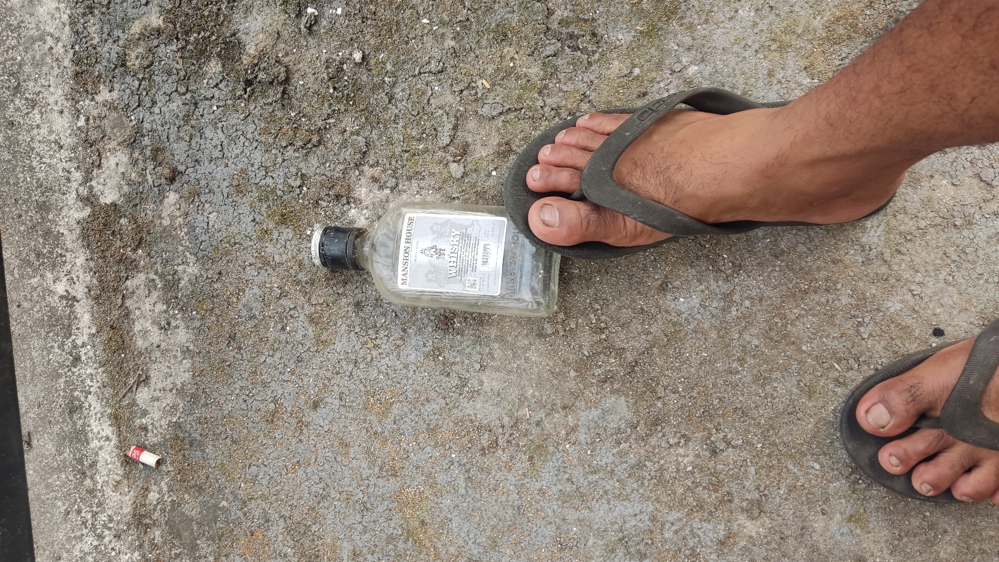 In Ngadiharjo, Indonesia, a bottle of Mansion House Whisky lies on a textured concrete surface. A person's right foot, clad in a dark flip-flop, rests on the bottle, obscuring part of its label. To the left of the bottle, a used cigarette butt is visible on the ground. The overall scene suggests a casual and possibly unkempt outdoor environment.