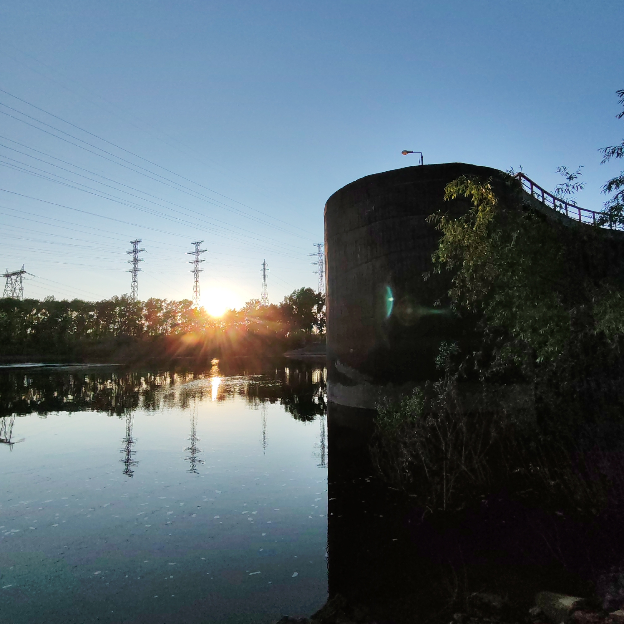 This image captures a serene sunset or sunrise over a body of water in Chaikovsky, Russia, blending natural beauty with industrial infrastructure. The sun, low on the horizon, casts brilliant golden rays that pierce through a line of trees on the far bank, creating a dramatic visual effect. Its light reflects intensely on the calm water surface, illuminating ripples and creating a bright path.
Several large power transmission towers stand prominently on the far bank, their intricate metal structures silhouetted against the bright sky. Numerous power lines stretch horizontally across the frame, converging towards the horizon. The reflections of these towers and lines are clearly visible in the tranquil water, adding depth to the scene.
On the right side of the foreground, a large, dark, curved concrete structure rises from the water. This robust piece of infrastructure, possibly part of a bridge or a dam, features a lamppost silhouetted against the sky on its top, along with a metal railing that follows its curve. Lush green foliage and bushes cling to the base and sides of this structure. A noticeable green lens flare is present in the mid-right of the image, a common photographic artifact when shooting into a bright light source.
The sky is a clear, pale blue, hinting at fair weather. The overall atmosphere is peaceful, combining the raw power of the sun and human-made structures with the quiet calm of the water. No people or visible text are present in the image.