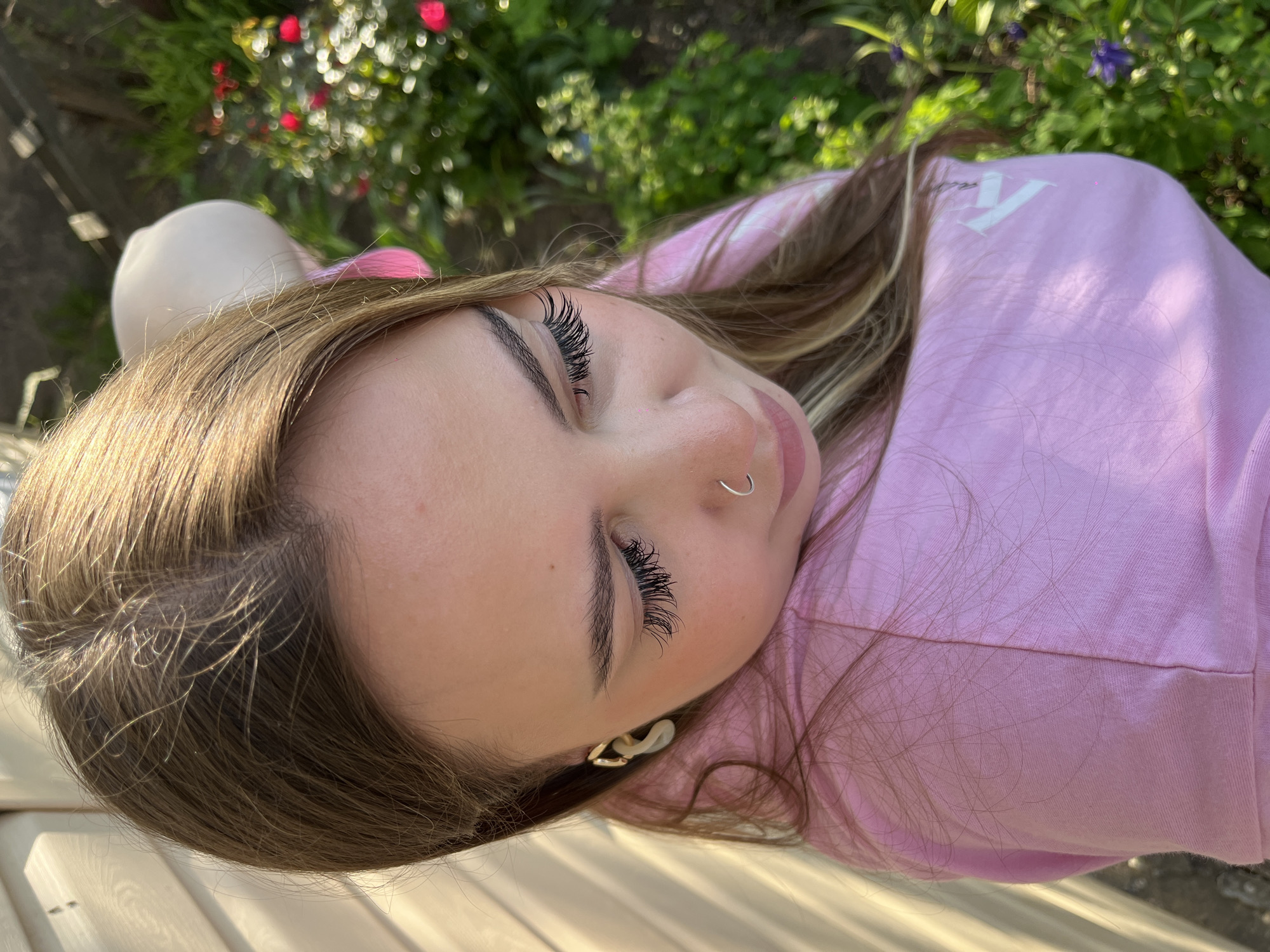 A young woman lies on a wooden surface outdoors, possibly a bench. She is wearing a light pink top and has brown hair with blonde highlights. Her eyes are closed, and she has long, dark eyelashes and a nose ring. The background includes greenery, including flowers, potentially suggesting a garden or park setting. The specific location is Novopokrovskaya, Russia.