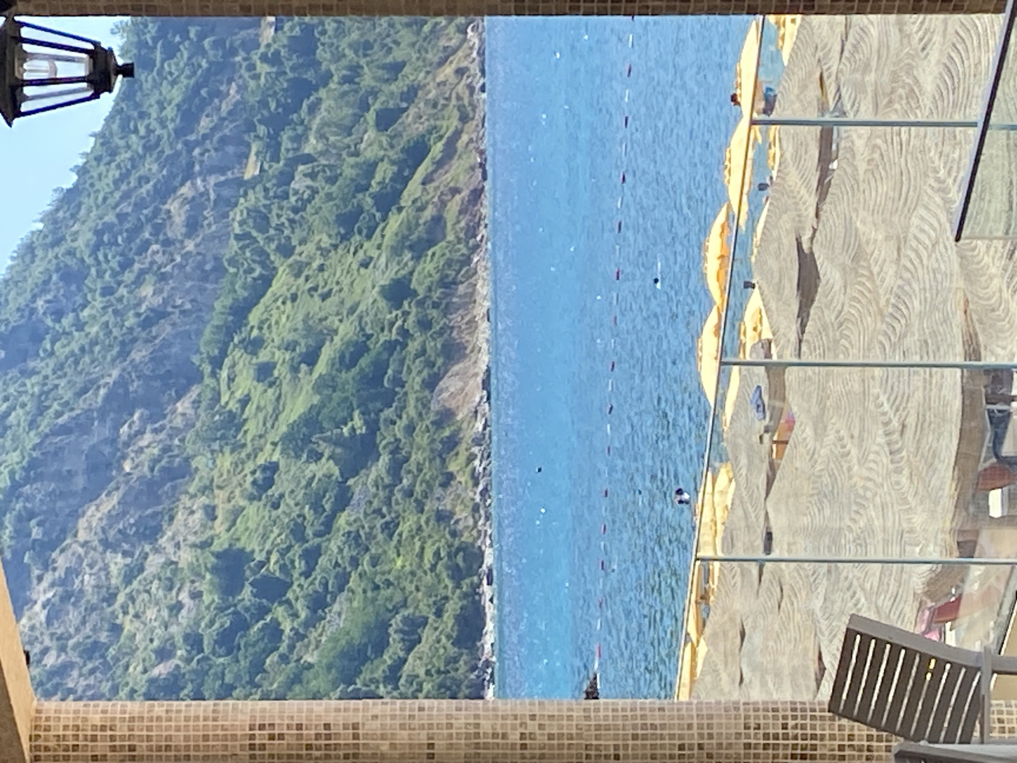 The image depicts a serene coastal scene, likely in Prijevor, Montenegro, as suggested by the context. The foreground is dominated by a tiled pillar on the left and a portion of a building facade on the right, framing a view of a beach and the sea. A glass barrier with metal supports is visible in the middle ground, separating the viewer's vantage point from the beach.
The beach itself is covered with numerous straw umbrellas, creating a textured pattern across the sand. A few beach chairs are also visible. In the distance, the bright blue sea stretches to the horizon, with a gentle swell and some buoys marking a swimming area. A few individuals can be seen in the water, appearing to be swimming or wading.
Beyond the sea, a lush green mountainside rises steeply, covered in dense vegetation and rocky outcrops. The sky is a clear, bright blue, indicating fair weather. A traditional-looking lantern hangs from the building on the right, adding a touch of architectural detail to the scene. The overall impression is one of a peaceful, sunny day at a popular seaside resort.