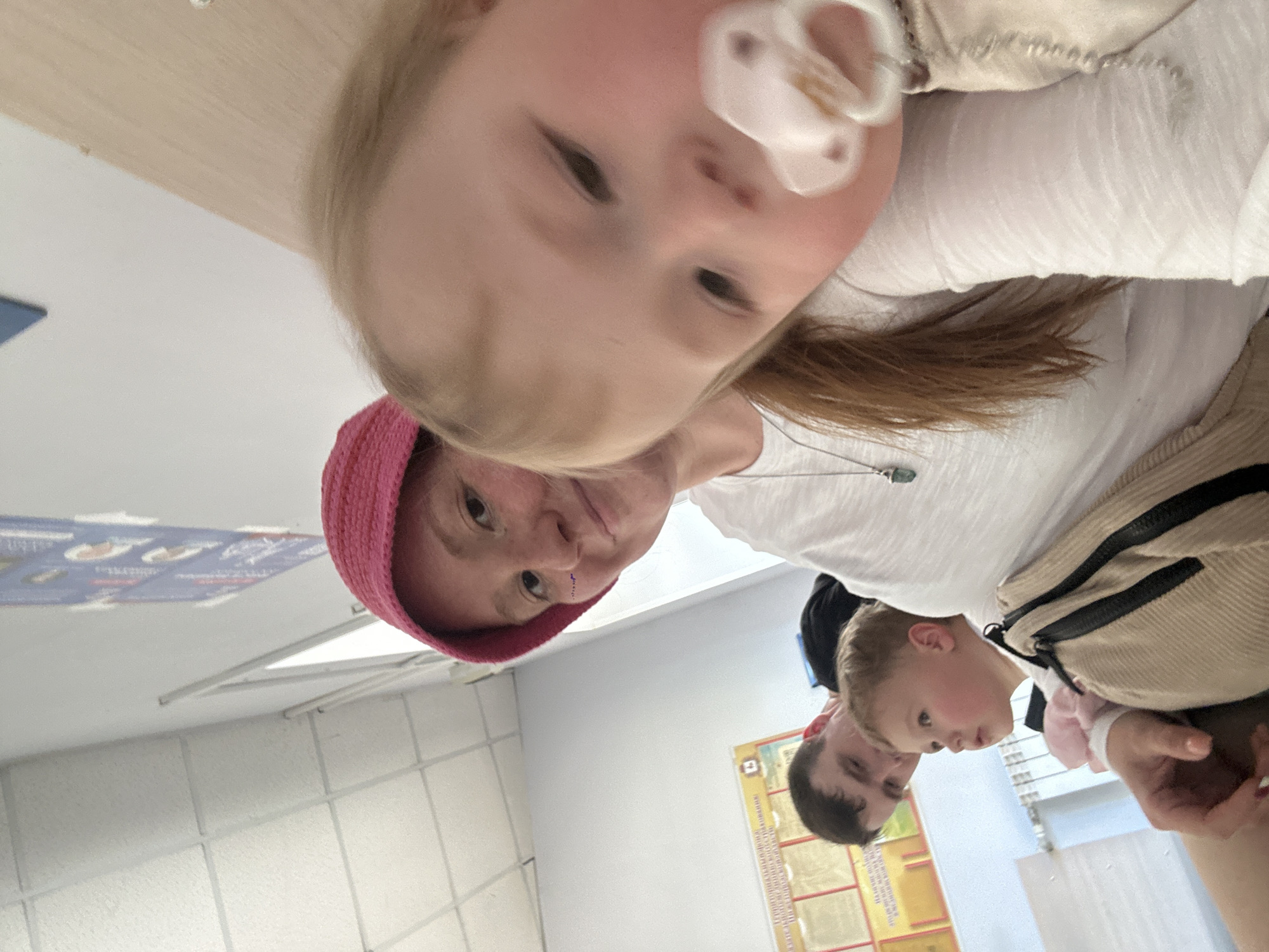 An indoor scene, taken from a low-angle selfie perspective, depicts a family in what appears to be a public waiting area or administrative office in Nizhny Novgorod, Russia.
The ceiling consists of white square acoustic drop tiles with visible seams. On the left wall, several framed documents or informational posters with Cyrillic text and diagrams are displayed. Below these posters, a white horizontal heating radiator is mounted against a light blue-grey wall. The room transitions to a plain white wall and then a light beige/wood-paneled wall on the right. A tall, narrow white vertical panel, possibly a room divider or whiteboard, is visible in the background, center-right.
In the immediate foreground, occupying the right side of the frame, is a blonde-haired baby or toddler with fair skin and rosy cheeks. The child wears a white long-sleeved top and has a white pacifier in its mouth, with eyes directed slightly downward and to the left.
Behind and to the left of the baby is an adult female, likely the mother, with reddish-brown hair visible below a bright pink crocheted bucket hat. She wears a white textured long-sleeved top and a silver necklace with a green pendant. Small purple dots or decorative stickers are applied to her right cheek as she smiles faintly towards the camera. Her left arm is visible, supporting or holding the youngest child.
Further left, a young child, approximately 2-4 years old, with light hair, is seated on the lap of an adult male. This child wears a light-colored top under a beige striped garment, possibly a small backpack or vest, and has a pink scrunchie or wristband on their right arm. The child looks towards the left, away from the camera.
The adult male, positioned in the background on the left, has dark, short-cropped hair and wears a dark shirt. He is partially obscured by the younger child and appears to be looking down past them. The overall scene suggests a family waiting together in an institutional setting.