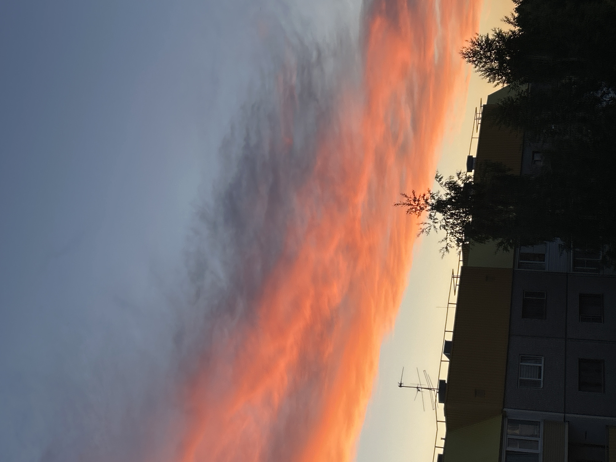 The image captures a striking view of a sunset or sunrise over a residential area, likely in Марковский, Russia. A dramatic, elongated band of clouds stretches horizontally across the sky, intensely illuminated with vibrant orange, red, and golden-yellow tones by the low sun. The sky above and below this radiant cloud formation is a softer, clear blue-grey.
In the foreground, on the right side of the frame, stands a multi-story apartment building with a facade featuring sections of dark grey and a lighter yellowish-brown. Rectangular windows are visible on its side. A distinct metallic television antenna with several horizontal rods is mounted on the building's roof or upper level. Silhouetted trees with sparse foliage partially obscure the building and rise against the bright horizon, notably towards the top right corner. The scene is devoid of people or discernible activity, highlighting the serene yet powerful display of nature's colors at dawn or dusk.
