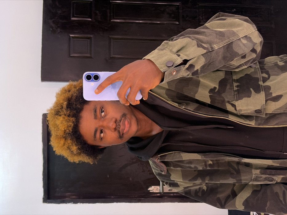 A Black male subject is depicted in an indoor environment in Igboefon, Nigeria, captured via a mirror selfie. The subject has dark skin, a short mustache, and voluminous, curly hair with yellowish-orange highlights on the top and dark roots. He is wearing a black hooded sweatshirt underneath an unzipped, green and dark green camouflage-patterned corduroy jacket featuring a brass-colored zipper and black buttons on its pockets and cuffs. The subject is holding a light purple smartphone with dual rear cameras in his right hand, angled to capture his reflection. His head is turned slightly, and his gaze is directed towards the viewer's left. The reflected background consists of a white wall segment on the left and a dark, paneled door with a visible silver-colored handle or hinge on the right. The scene is uniformly lit.