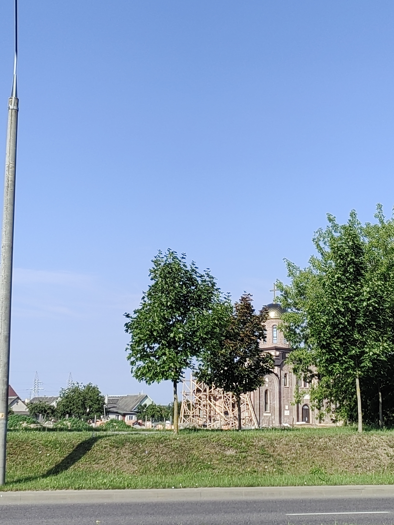 This is a daytime, eye-level, full-shot photograph taken from the side of a road in Hrodna, Belarus. In the foreground, a paved road runs horizontally across the bottom of the frame. Behind the road is a strip of grassy embankment. In the middle ground, there is a row of trees, with a church under construction visible behind them. The church has a dome with a cross and is made of brick. To the left of the church, there are several residential buildings and what appear to be power lines in the distance. The sky is clear and blue. A tall, slender, grey pole is visible on the left side of the image, extending from the bottom to the top of the frame.