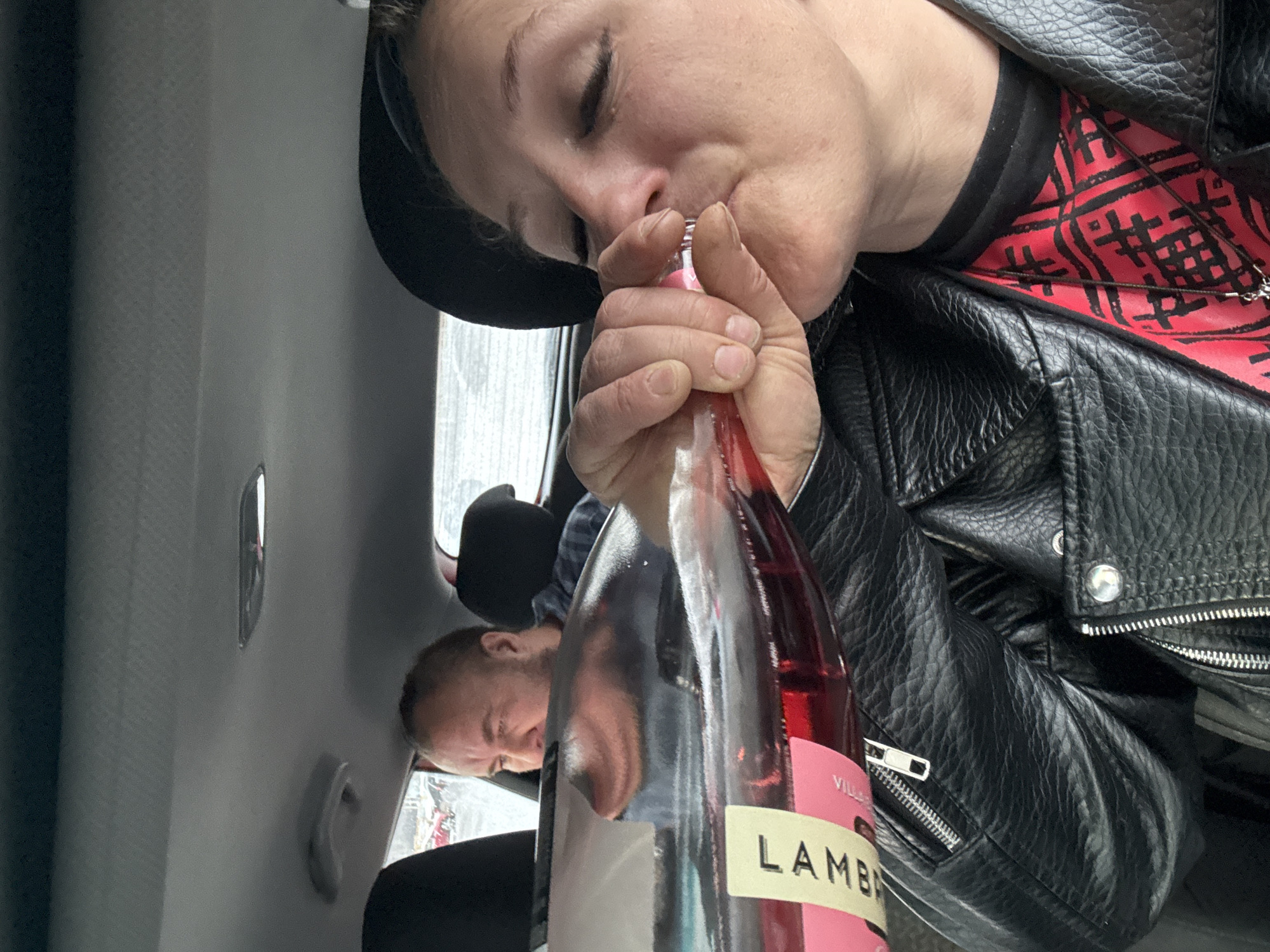 The image shows a woman in a car drinking directly from a bottle of LAMBRUSCO wine. She is wearing a black hat and a black leather jacket over a red and black patterned shirt. A man is visible in the background, seated behind her. The interior of the car is grey. No specific details in the image confirm the location as Izhevsk, Russia.